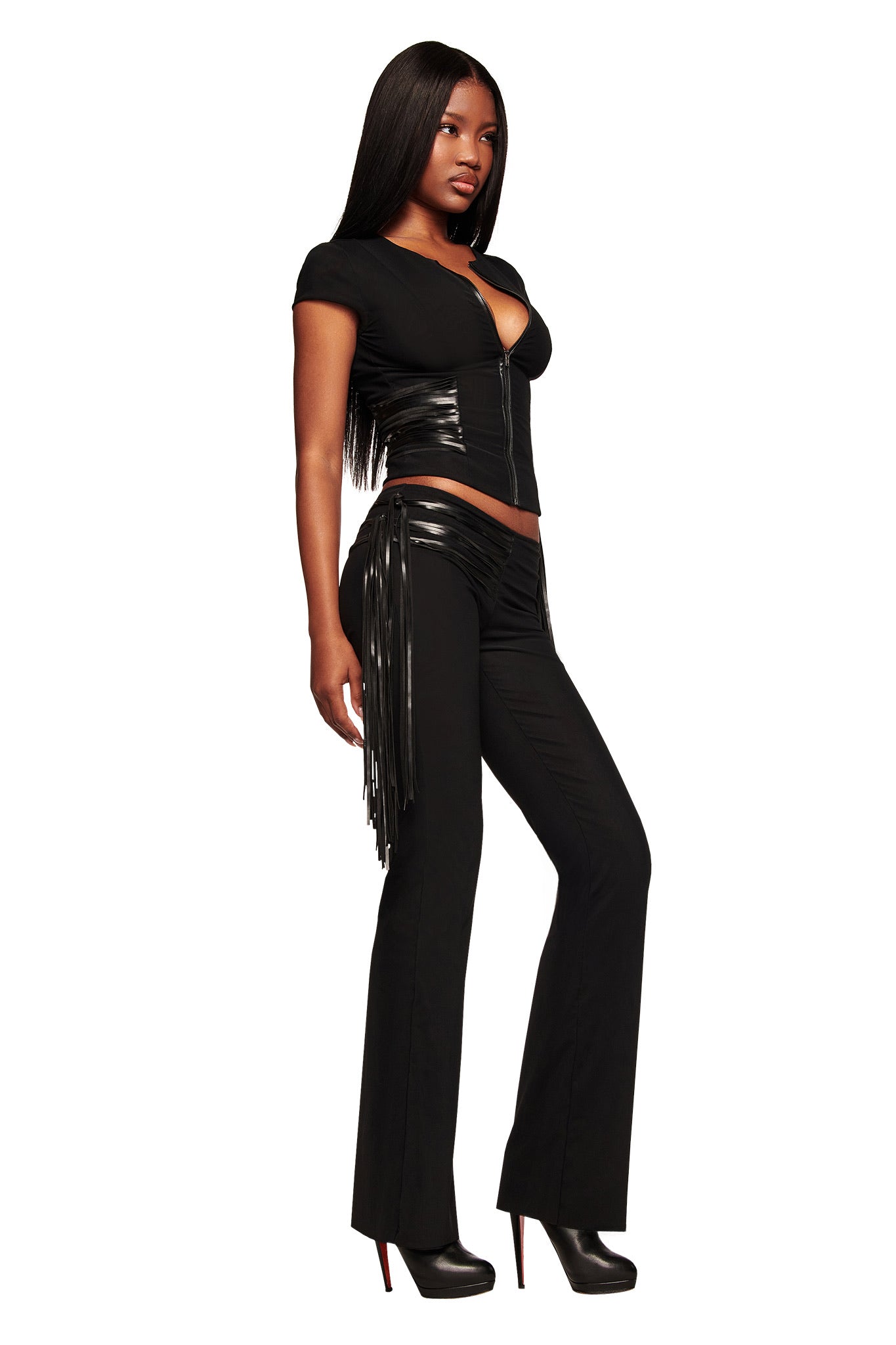 A woman poses confidently against a white background in the ISELDA FRINGE PANT - BLACK by I.AM.GIA, featuring fitted black pants with shiny faux leather fringe on the sides, paired with a short-sleeved, zip-up top with shiny accents and black high heels.