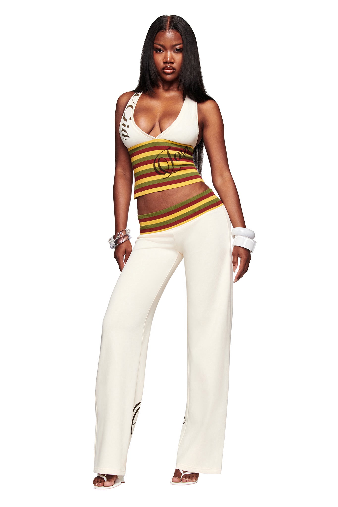 A woman stands confidently in the I.AM.GIA CANDI TOP in cream—a V-neck racer back with green, yellow, and red stripes—paired with white wide-leg pants, bracelets, high-heeled sandals, and long straight hair.