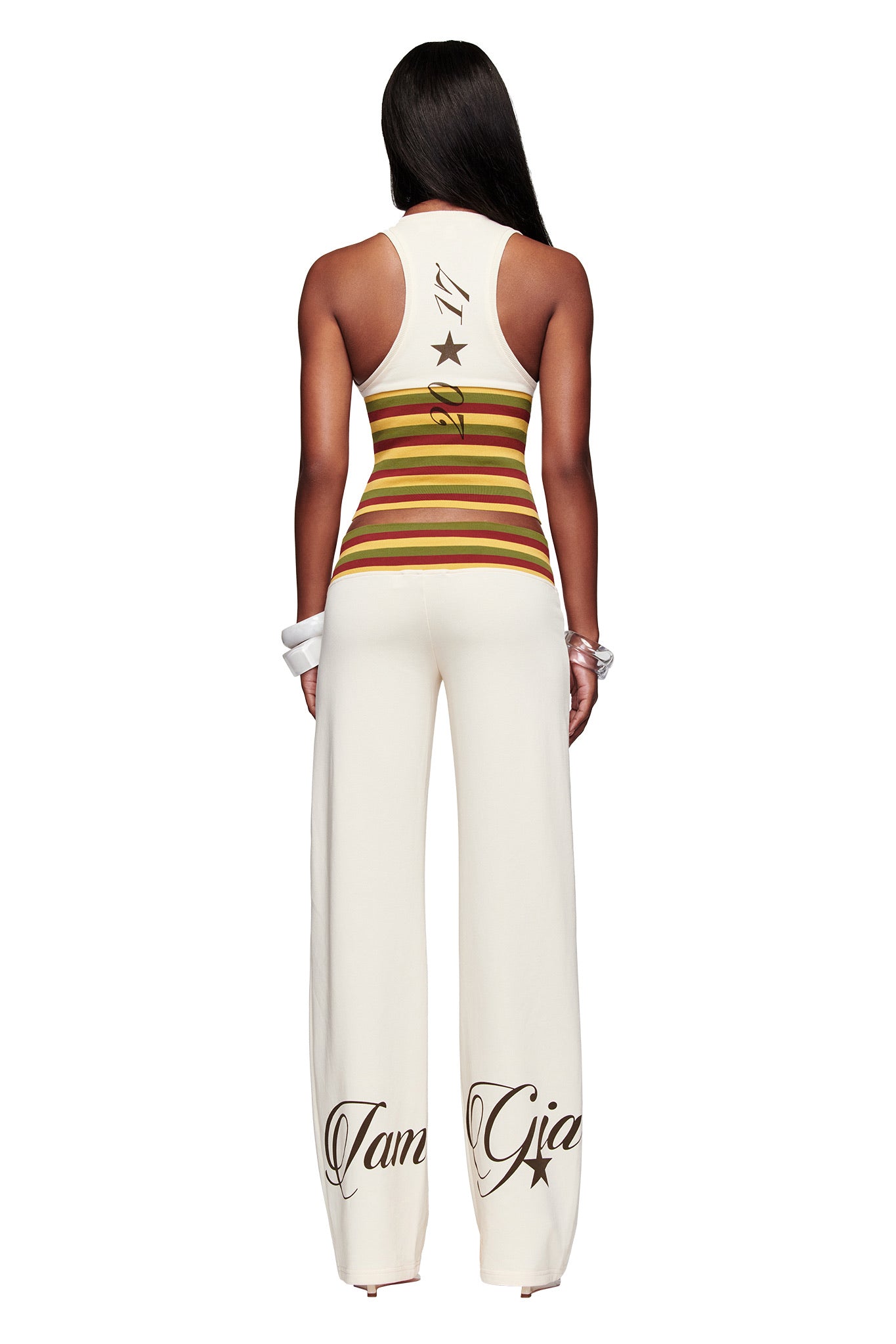 A person stands facing away, wearing a sleeveless striped top and I.AM.GIA CANDI PANT - CREAM wide leg tracksuit pants with bold script text near the ankles, set against a white background.