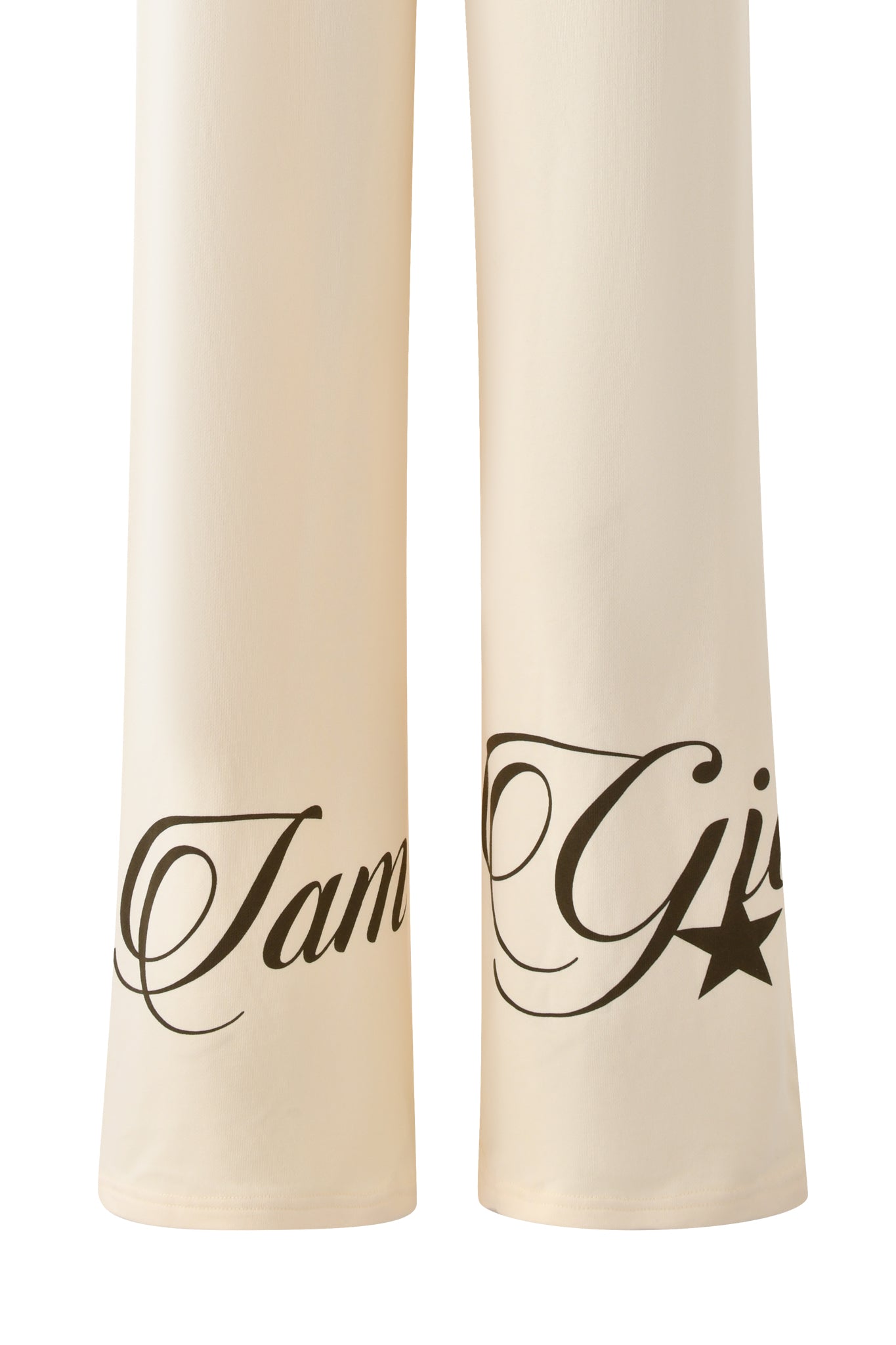 The image shows the lower part of the I.AM.GIA CANDI PANT in cream, featuring black cursive writing and a star near the hems. The visible “I am G” text hints at the signature I.AM.GIA tracksuit style.