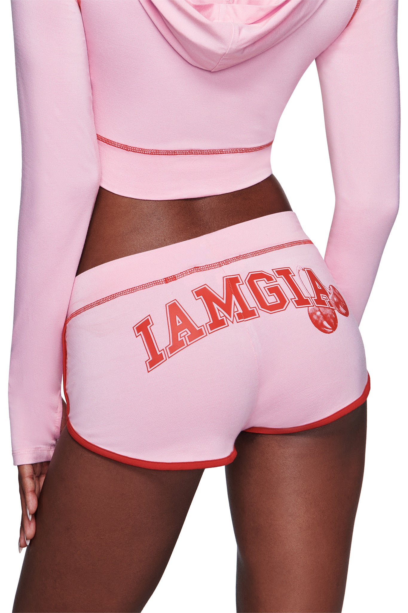 A person wearing a pink hoodie and the I.AM.GIA BLARE DOLPHIN SHORT in baby pink, featuring red trim and bold red "IAMGIA" text on the back. The person's hand rests at their side.