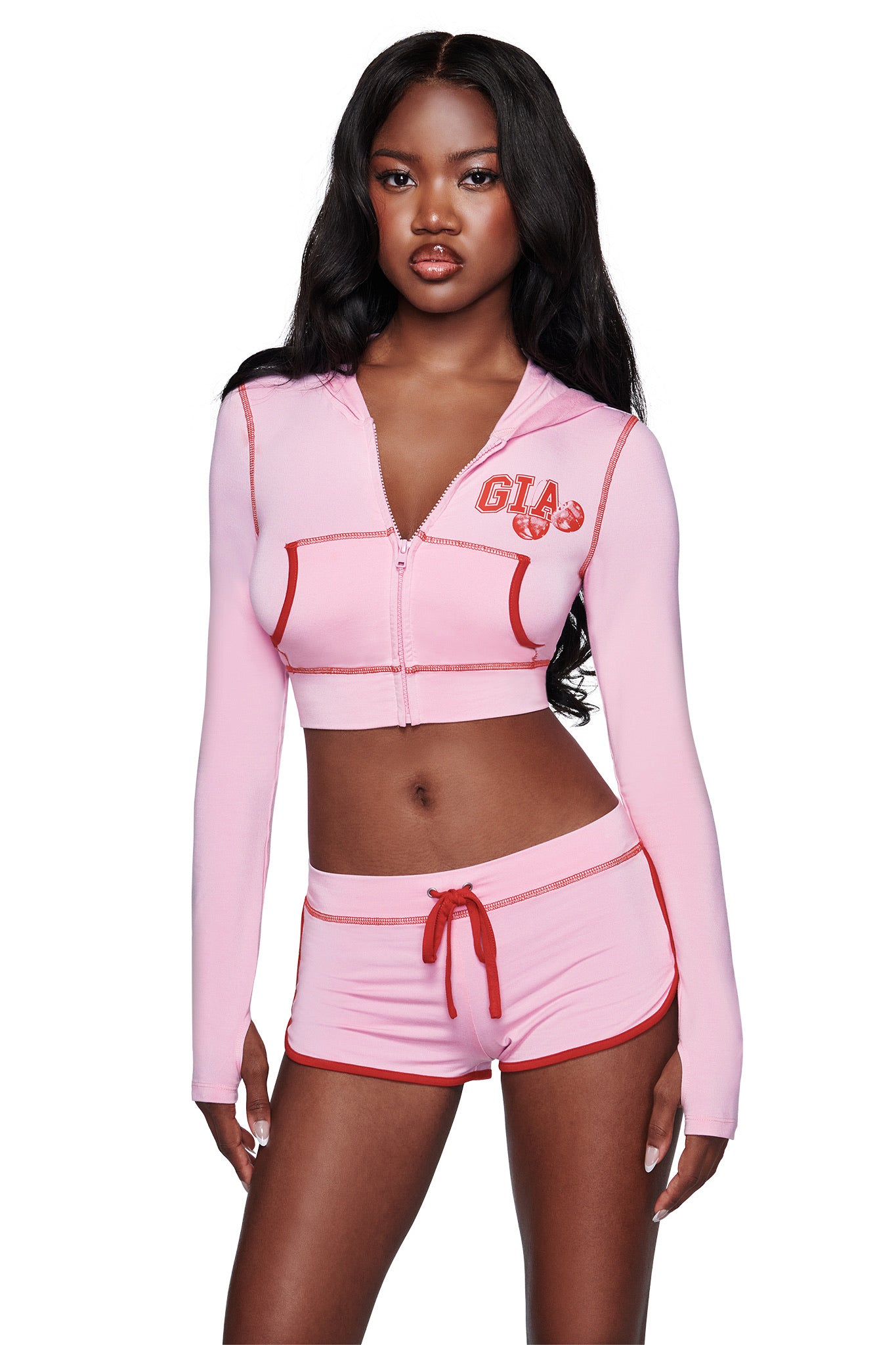 A woman with long black hair wears a pink cropped zip-up hoodie and matching I.AM.GIA BLARE DOLPHIN SHORT - BABY PINK shorts with red trim, standing against a white background.