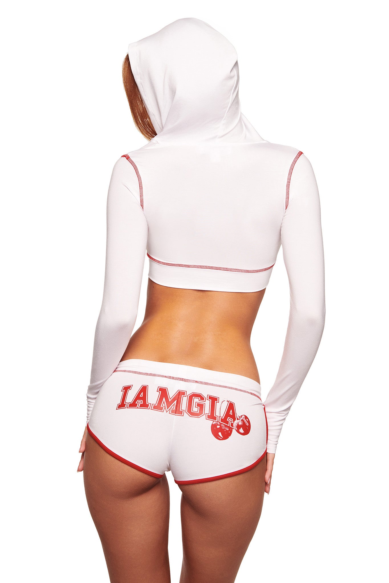 A woman stands with her back to the camera, wearing I.AM.GIA's BLARE DOLPHIN SHORT in white, paired with a white cropped hoodie featuring red trim and the bold I.AM.GIA logo in red.