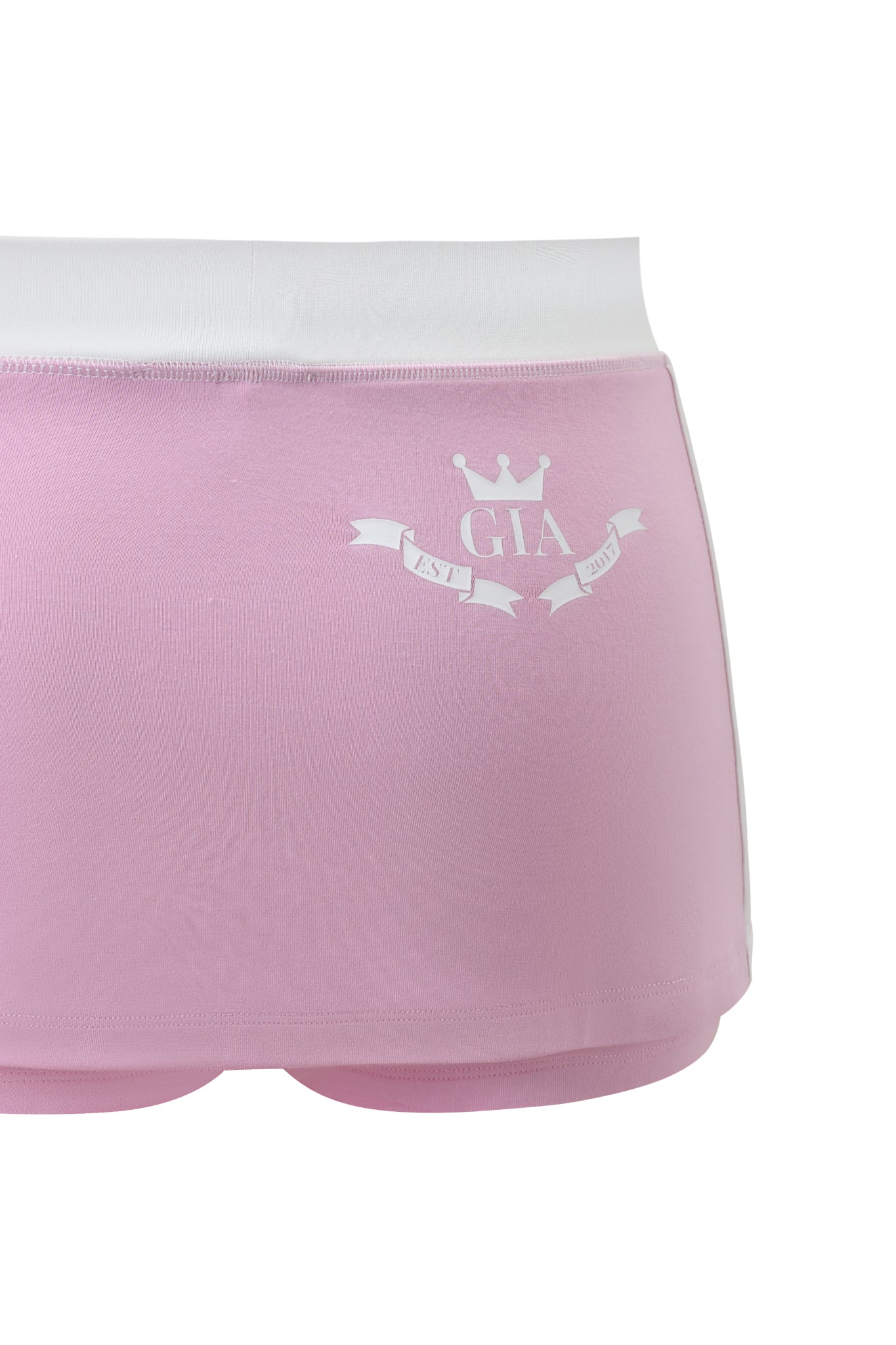 A close-up of the I.AM.GIA BLARE PIPING MICRO SKORT in baby pink with a white waistband. The back has a white crown graphic above "GIA" and a banner below reading "EST. 2007.