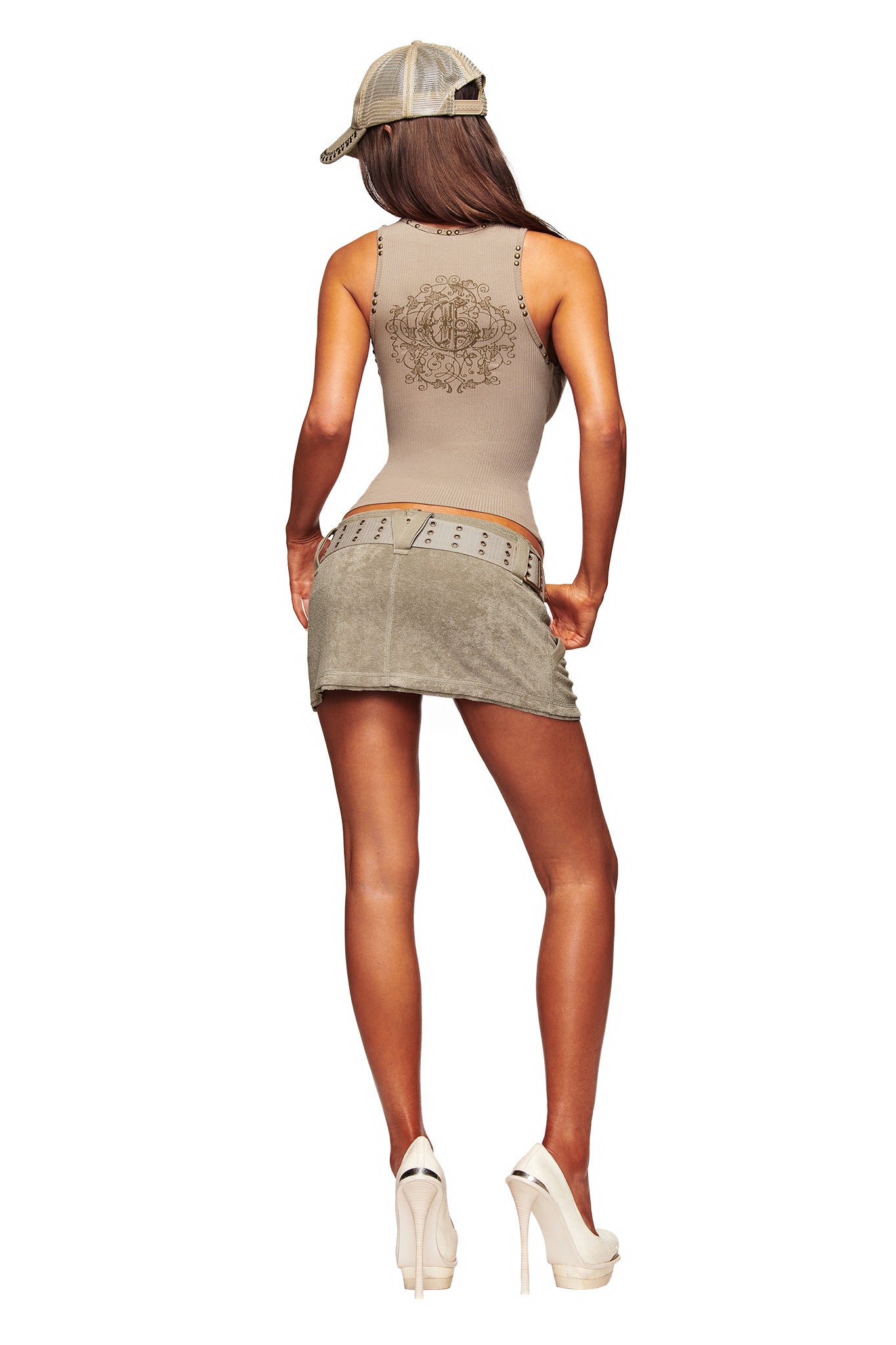 A woman poses with hands on hips in a beige tank top, ISSHA TERRY MINI SKIRT - STONE by I.AM.GIA with detachable canvas belt, a beige cap, and heels. She faces away, showing her long hair against a white background.