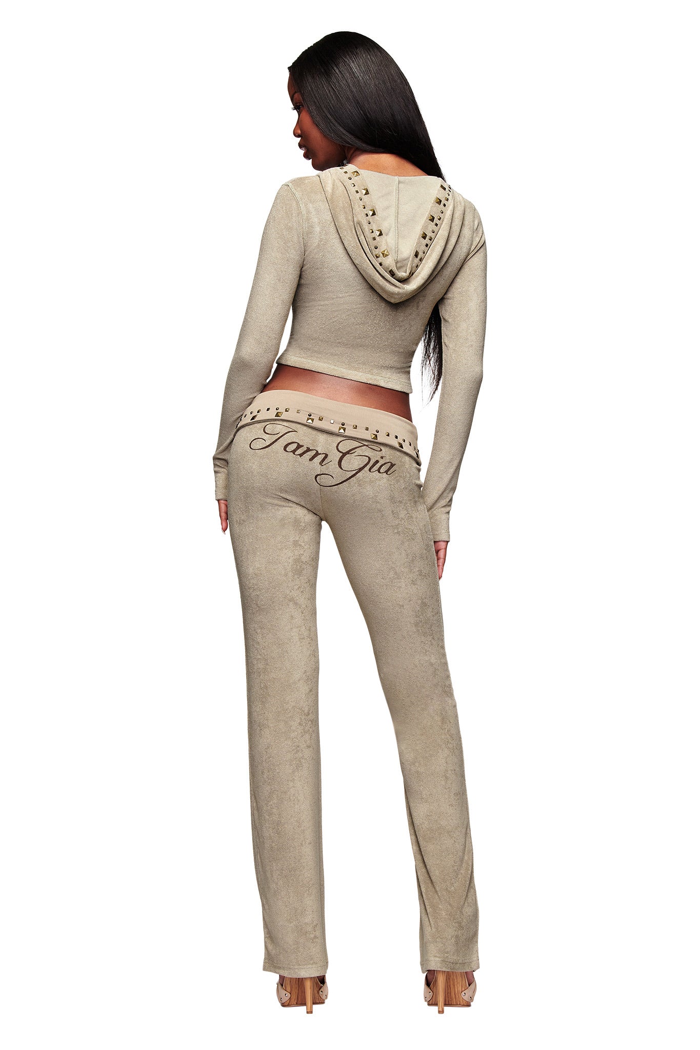 A woman stands facing away, wearing the I.AM.GIA BLARE STUDDED TERRY PANT in Stone. The back features rhinestones spelling “I Am Gia.” She pairs the fitted velour trackpant with a studded hooded top and high heels.