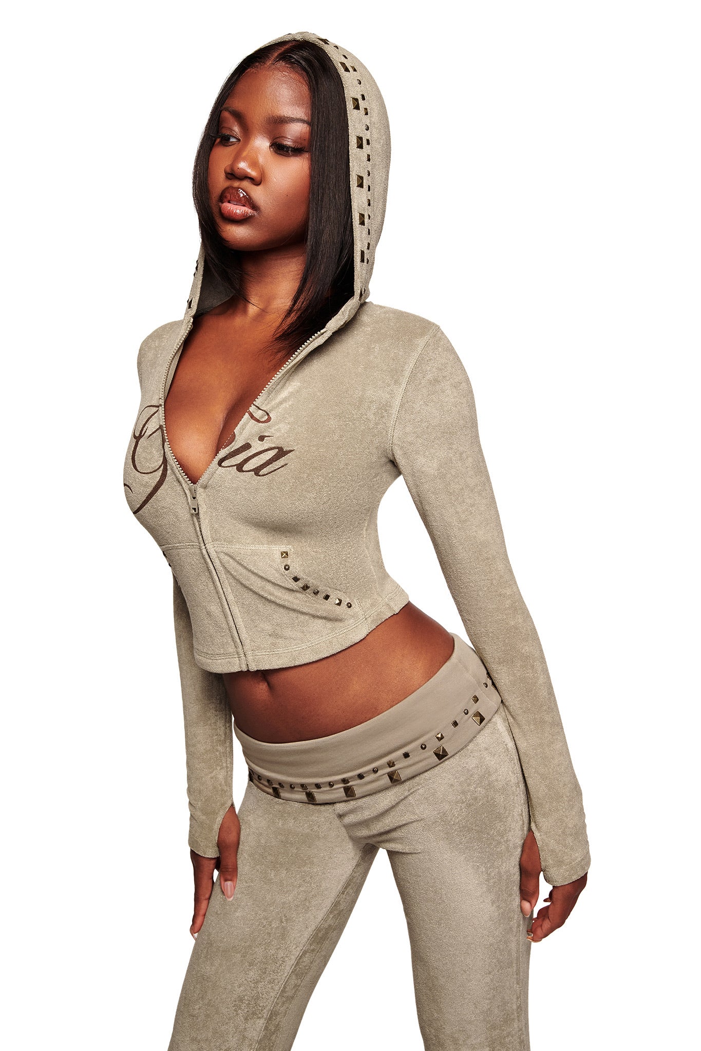 A woman in the I.AM.GIA BLARE STUDDED TERRY HOODIE in stone, featuring studded details and script lettering, poses against a white background, showcasing the brand's signature style.