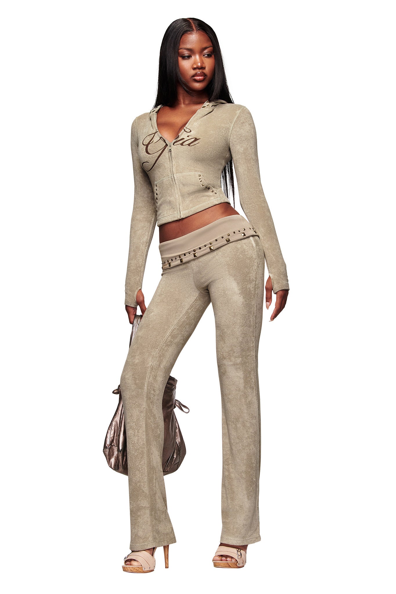 A woman with long straight hair wears I.AM.GIA's BLARE STUDDED TERRY PANT in stone, styled with a cropped zip-up hoodie and heels. She carries a shiny handbag and stands confidently against a white background.