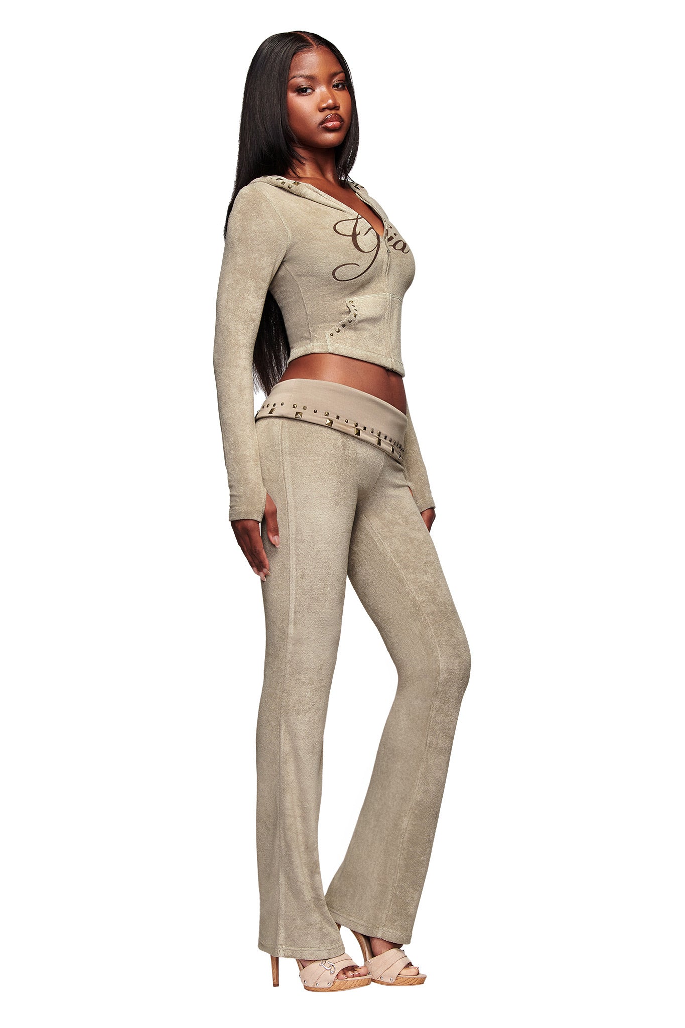 A woman poses confidently in a beige, fitted top with a lace-up neckline and the I.AM.GIA BLARE STUDDED TERRY PANT - STONE, styled with high-heeled sandals against a white background.