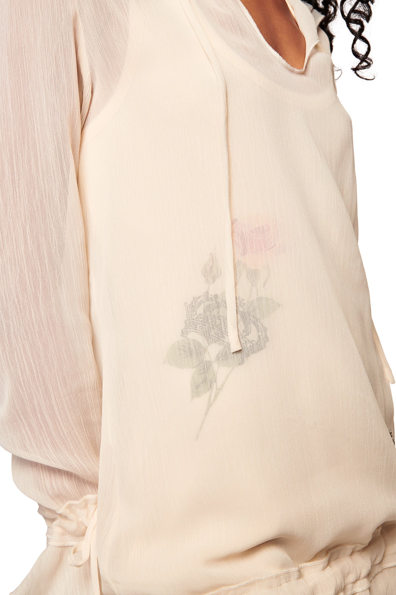 A close-up of a person wearing the I.AM.GIA DIAMOND 2-IN-1 MINI DRESS in cream, featuring sheer georgette fabric, long sleeves, and a subtle rose floral pattern visible near the chest.