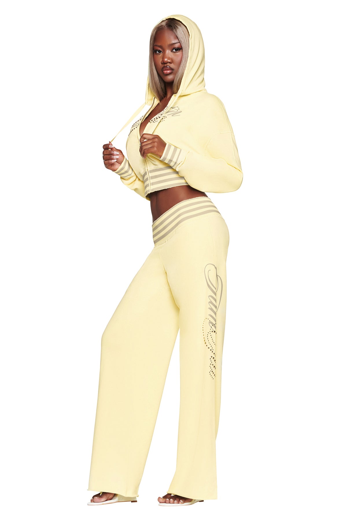A woman poses confidently against a white background wearing the I.AM.GIA VEDA JACKET in yellow—a cotton-blend fleece cropped zip-up hoodie paired with matching flared pants that feature decorative writing on the left leg.
