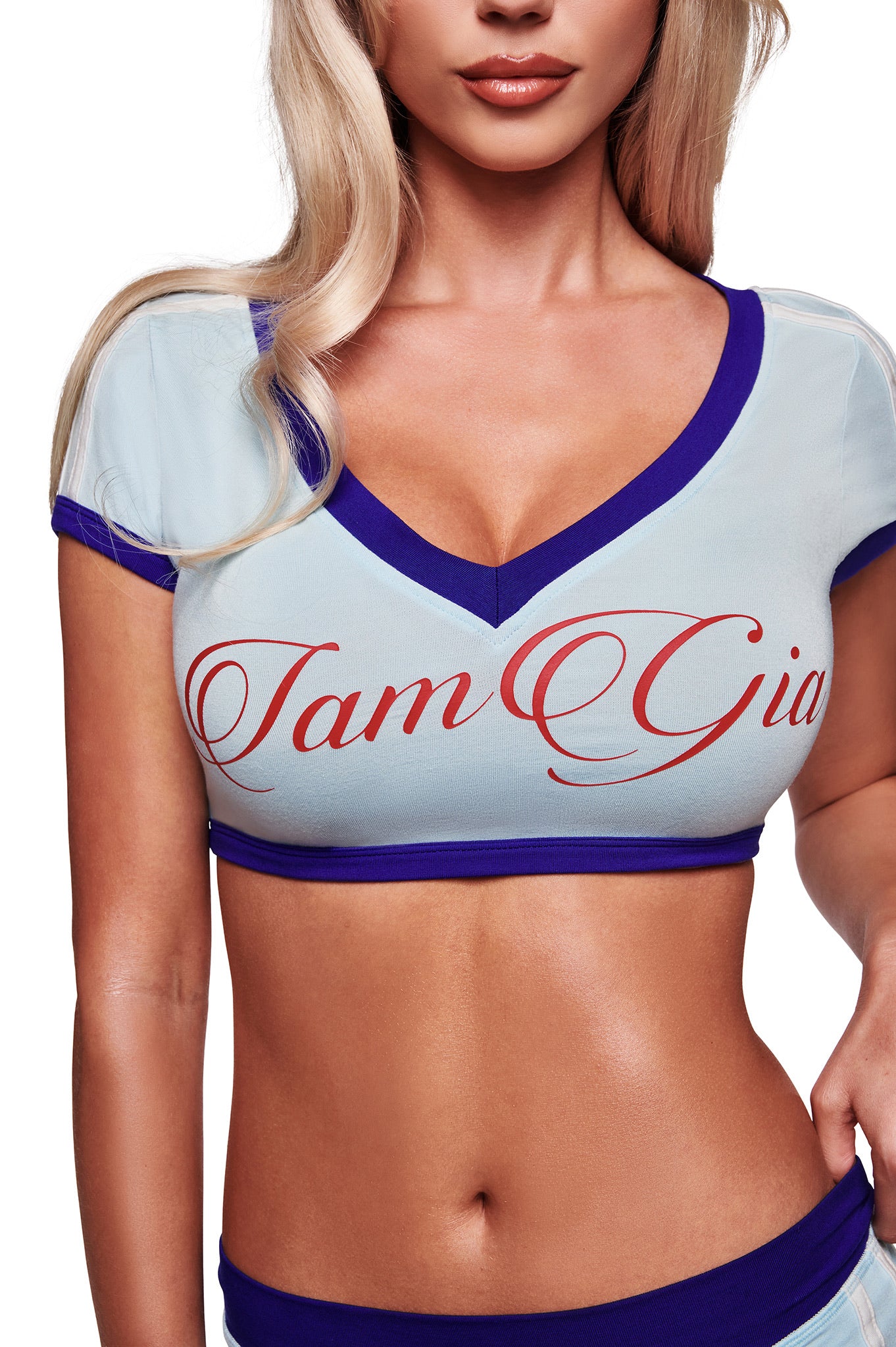 A woman wears the BLARE SPORT CROP TEE AND HEADBAND in baby blue by I.AM.GIA, featuring purple trim, a sporty v-neck, and bold red "I.AM.GIA" script. Her blonde hair falls over her shoulder as her face is partly visible.