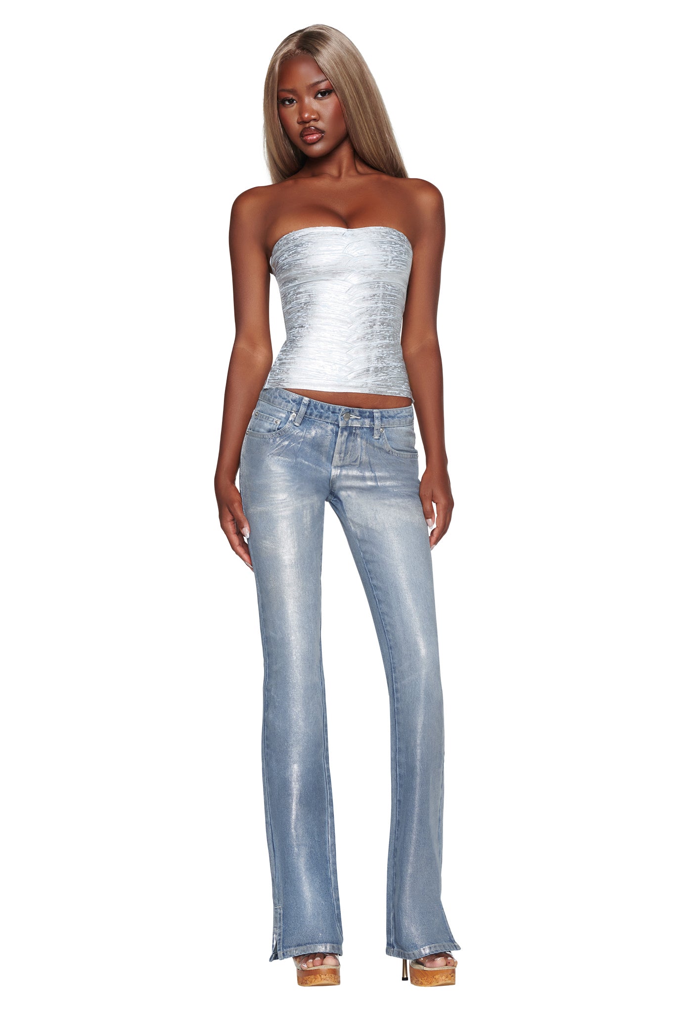 A woman with long, straight blonde hair models the I.AM.GIA KHIA BANDAGE BOOB TUBE in metallic blue, styled with light blue flared jeans and platform heels. She stands facing forward against a plain white background, her sleek ensemble highlighting a modern bodycon silhouette.