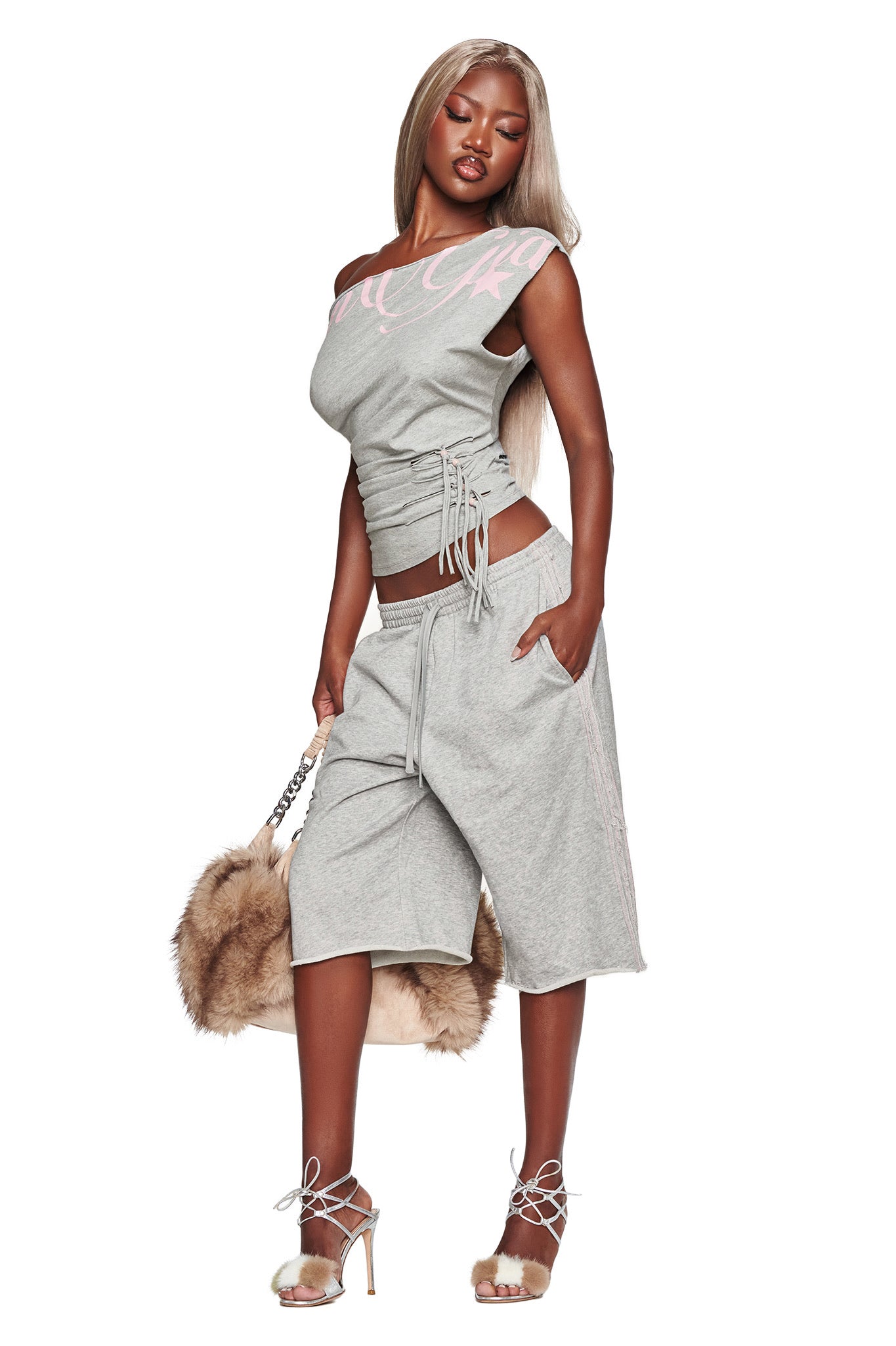 A woman wearing the I.AM.GIA BEX OFF SHOULDER TOP in grey poses with one hand in her pocket and holds a large faux fur bag. She pairs the top with matching long shorts, wears heels, and has long, straight hair. The background is plain white.