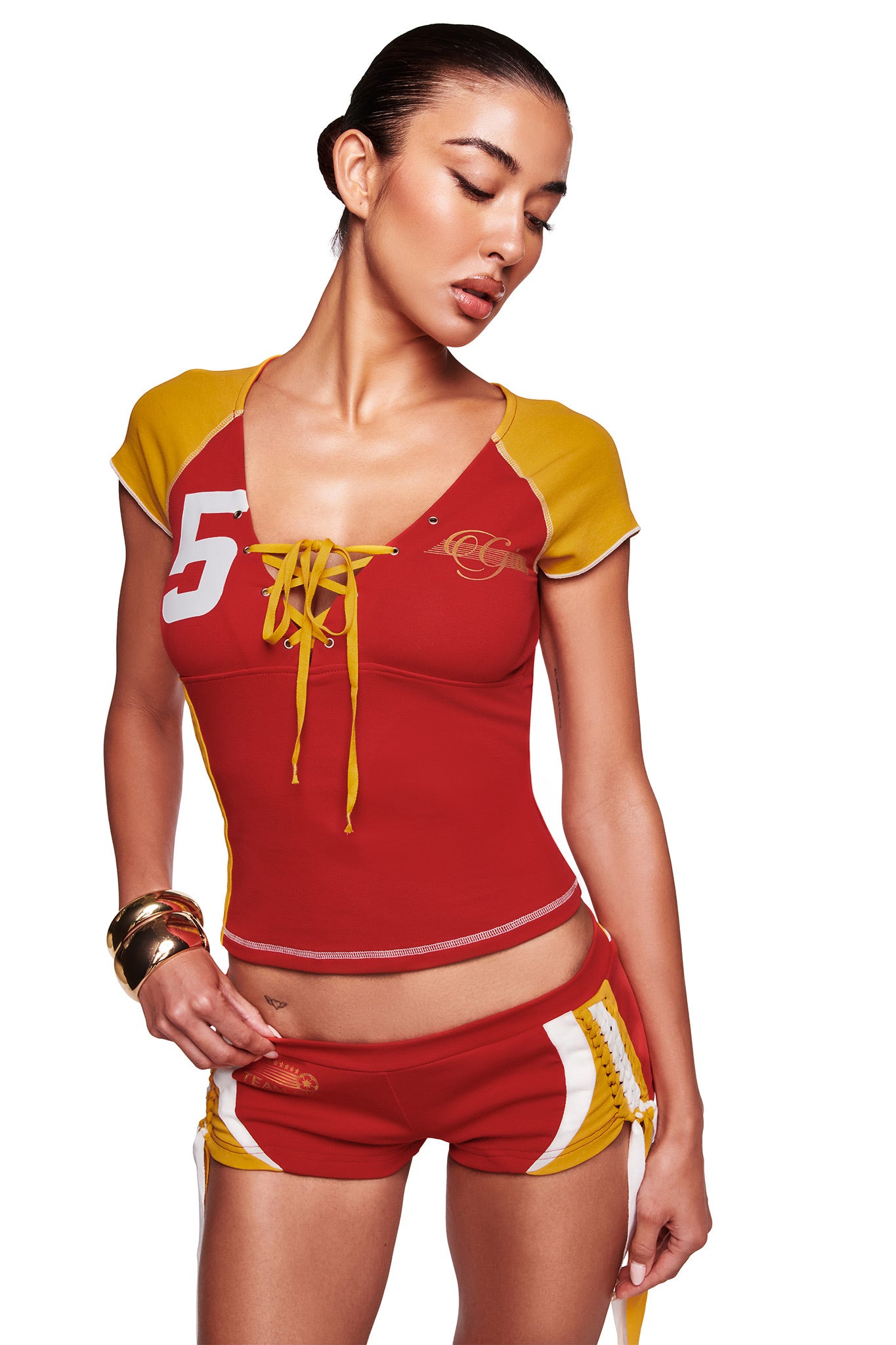 A woman models the I.AM.GIA NOVIA LACE-UP T-SHIRT AND HEADBAND in red, featuring a lace-up front and sporty design, paired with matching shorts and gold bangles against a white background.
