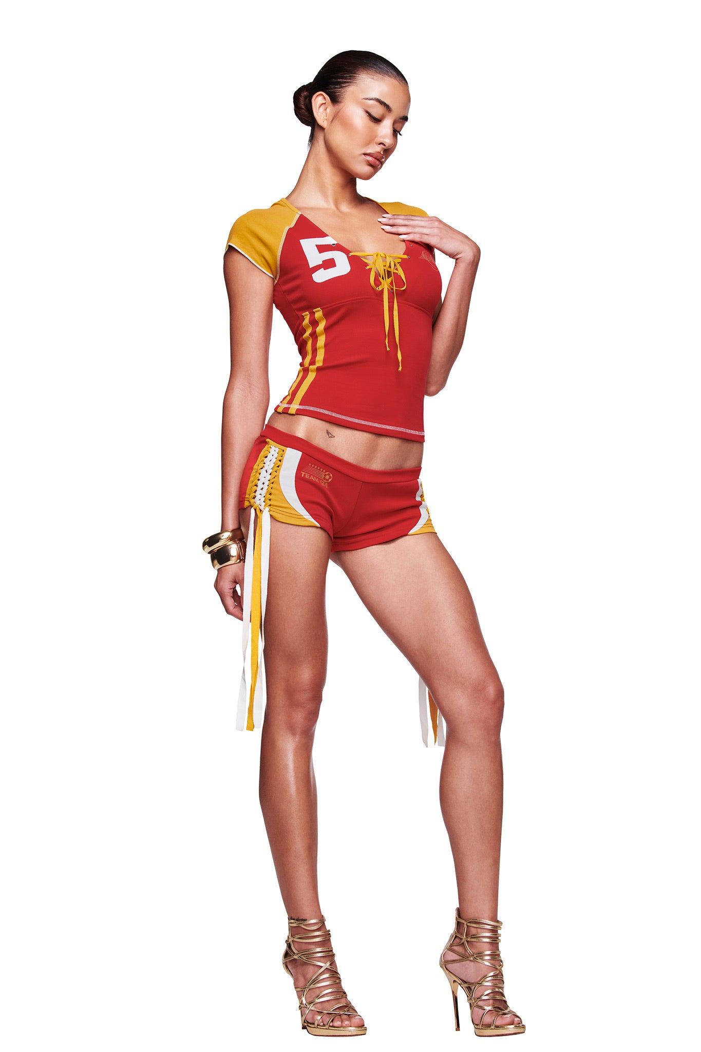 A woman poses against a white background in the I.AM.GIA NOVIA MICRO SPORT SHORT - RED and a matching sporty top with "5" and lace-up front, styled with gold high-heeled sandals.