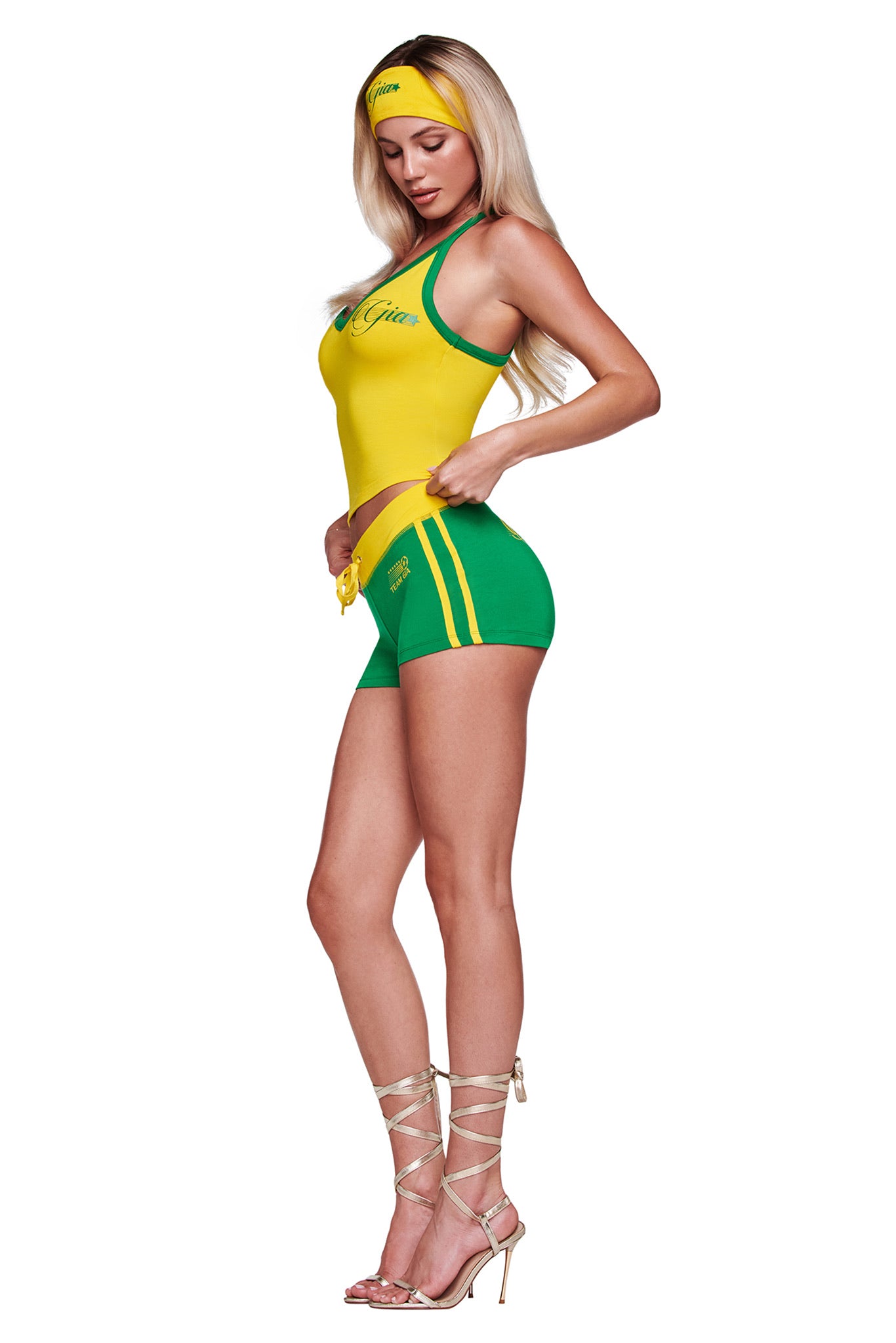A woman in profile adjusts her I.AM.GIA BLARE PIPING SHORT - GREEN, paired with a yellow tank top, yellow headband, and strappy high-heeled sandals. Her long blonde hair is loose.