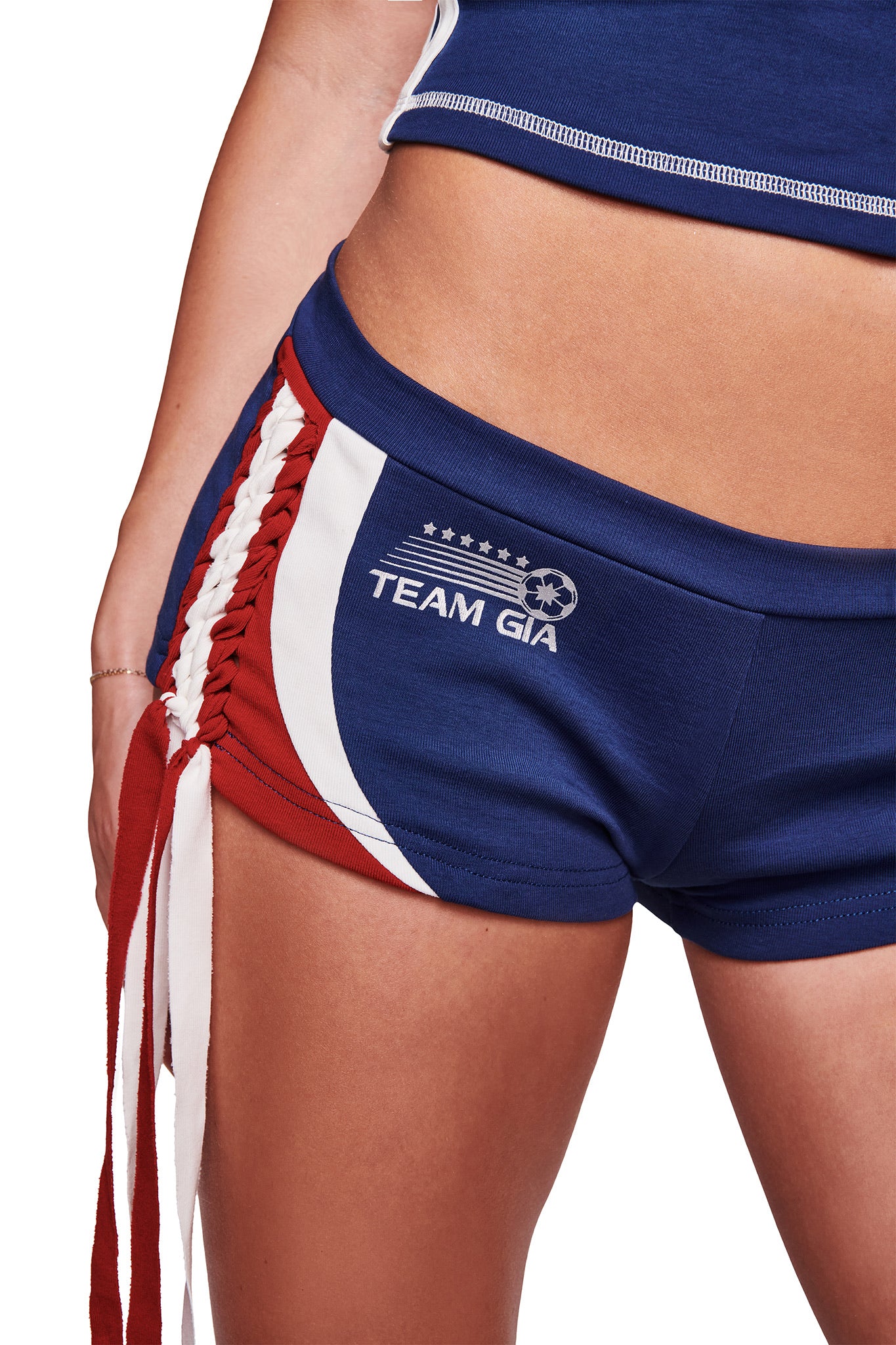 Close-up of a person wearing I.AM.GIA NOVIA MICRO SPORT SHORT - NAVY, featuring red and white side stripes, lace-up front, and "TEAM GIA" with stars and a soccer ball. Only the lower torso and one arm are visible.