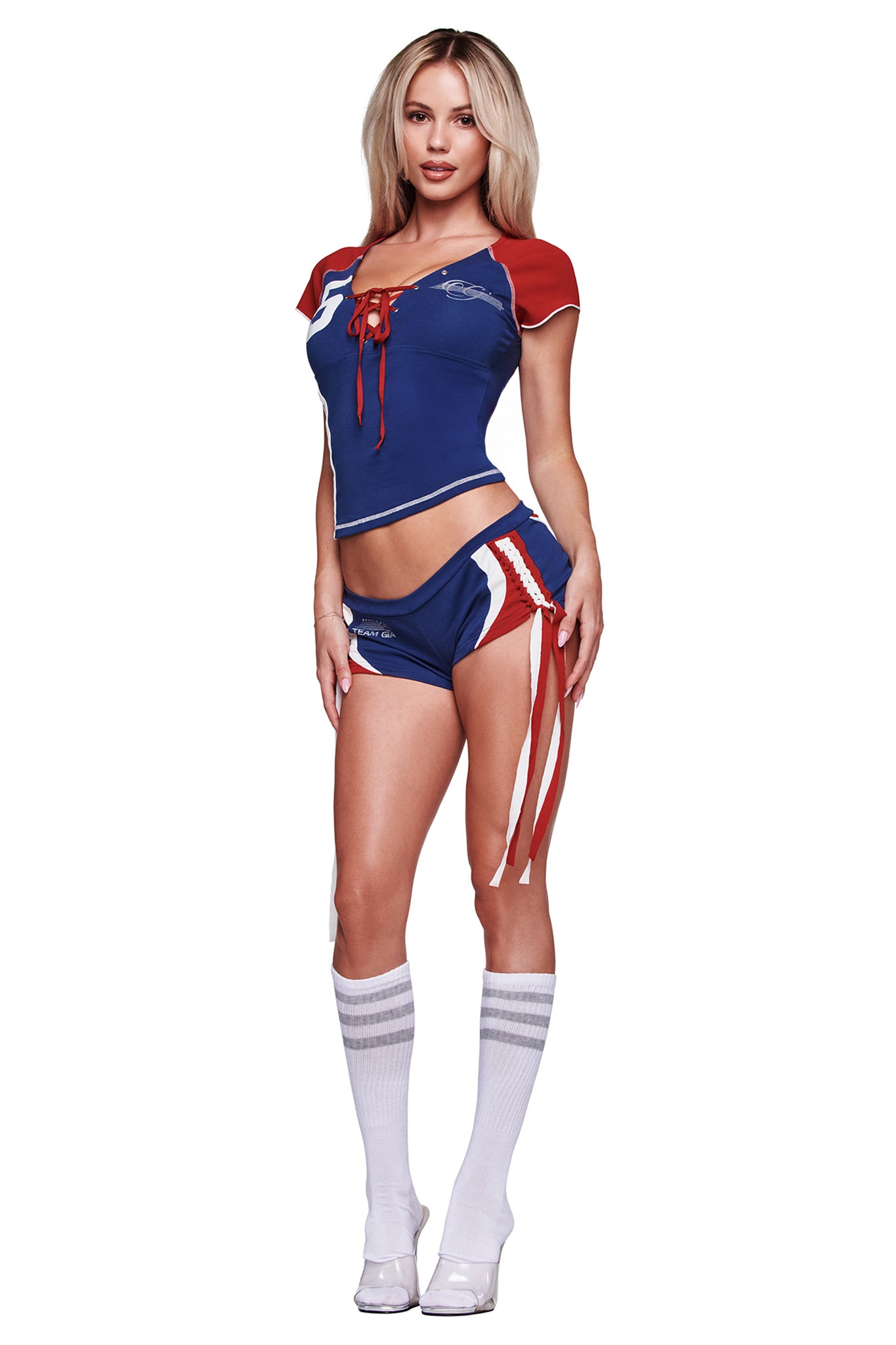A woman faces forward wearing I.AM.GIA's NOVIA MICRO SPORT SHORT - NAVY, a sporty stretch cotton short with side ties, paired with a blue, red, and white top, knee-high striped socks, high-heeled shoes, and long blonde hair.