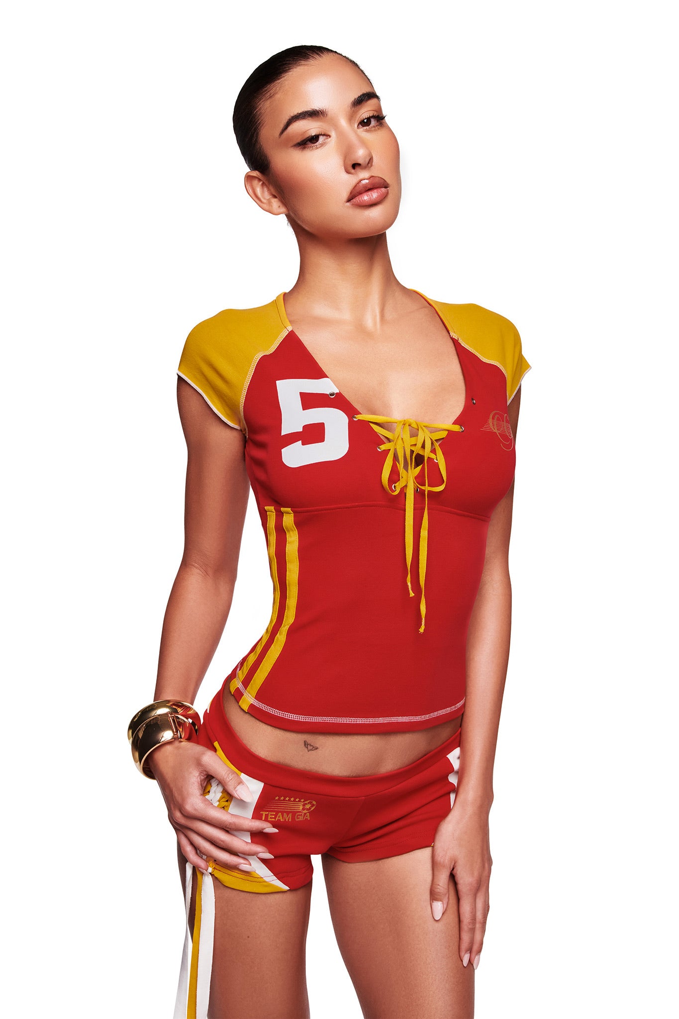 A woman poses confidently in I.AM.GIA's red NOVIA LACE-UP T-SHIRT AND HEADBAND, styled with gold bangles and slicked-back hair, against a white background.