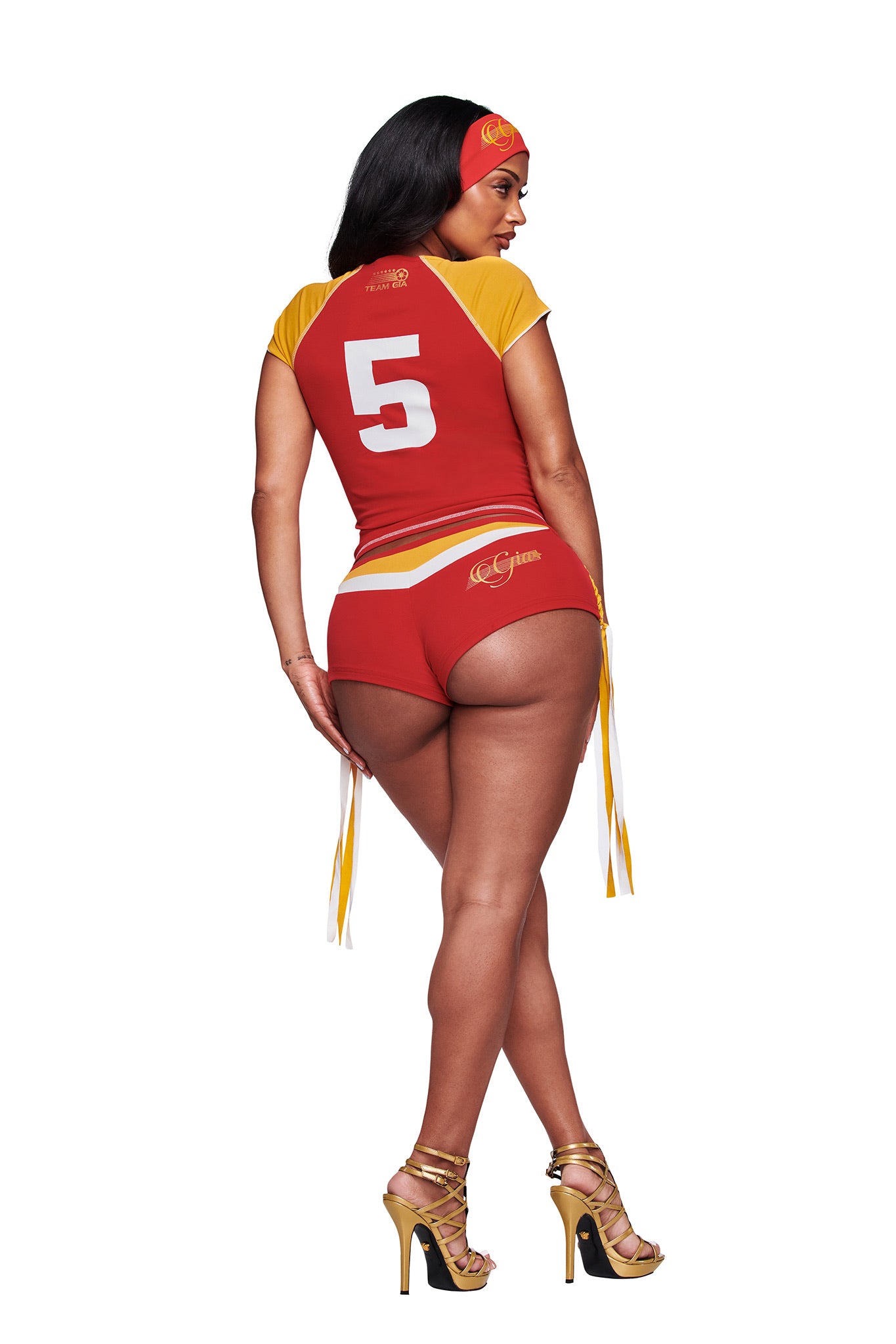 A woman stands against a white background, facing away in I.AM.GIA NOVIA MICRO SPORT SHORT - RED, part of a red and yellow sports outfit with the number 5 on the back, gold high heels, and a matching headband.