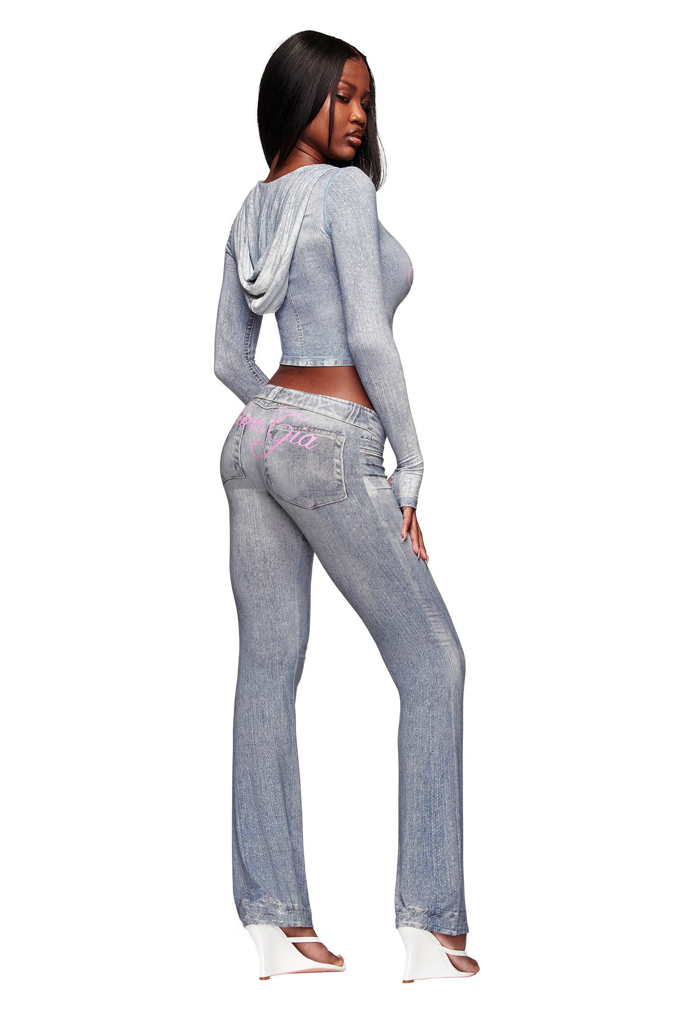 A woman poses in I.AM.GIA's BLARE TRACKPANT in Denim Trompe L'oeil, paired with a matching light blue faux-denim hooded top and white open-toe heels. She has long, straight black hair.