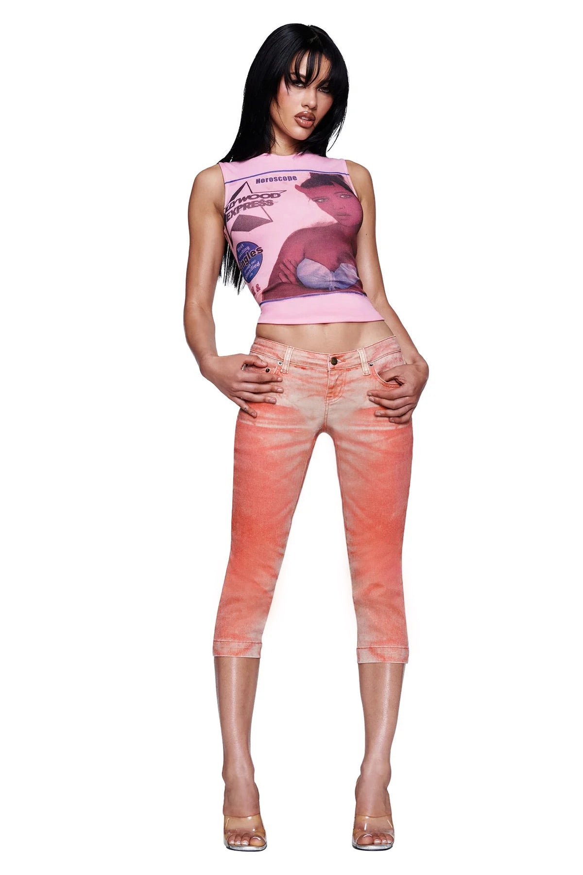 A woman wearing a pink sleeveless crop top with a graphic print, the I.AM.GIA SUNSET CAPRI JEAN in Peach ombré wash, and clear high-heeled shoes stands against a white background.