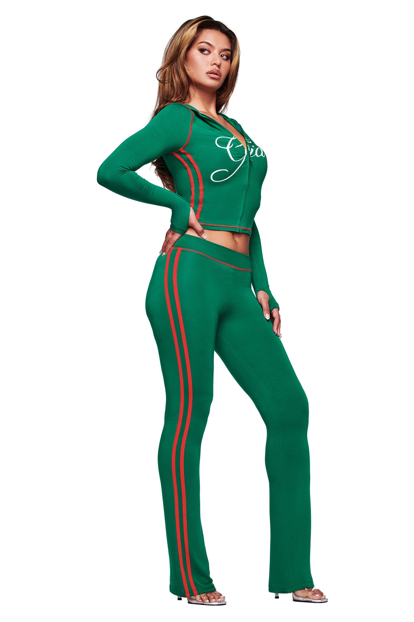A woman poses against a white background in the I.AM.GIA BLARE SPORT HOODIE AND HEADBAND - GREEN, styled with matching high-waisted pants and open-toed heels.
