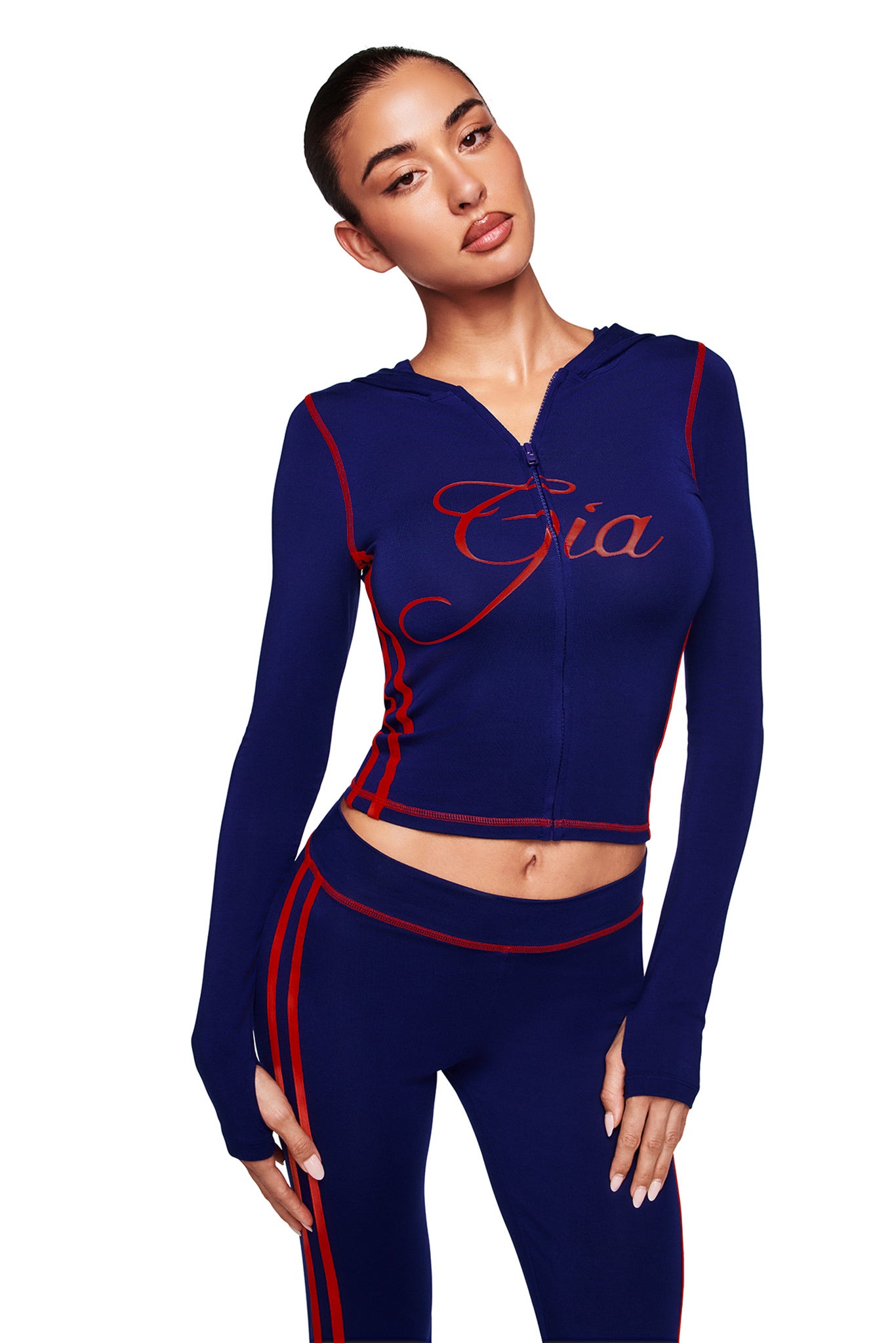 A woman wears the I.AM.GIA BLARE SPORT HOODIE AND HEADBAND in navy, featuring red piping and "Gia" script, paired with matching navy pants with red stripes and a bodycon fit, posing against a white background.