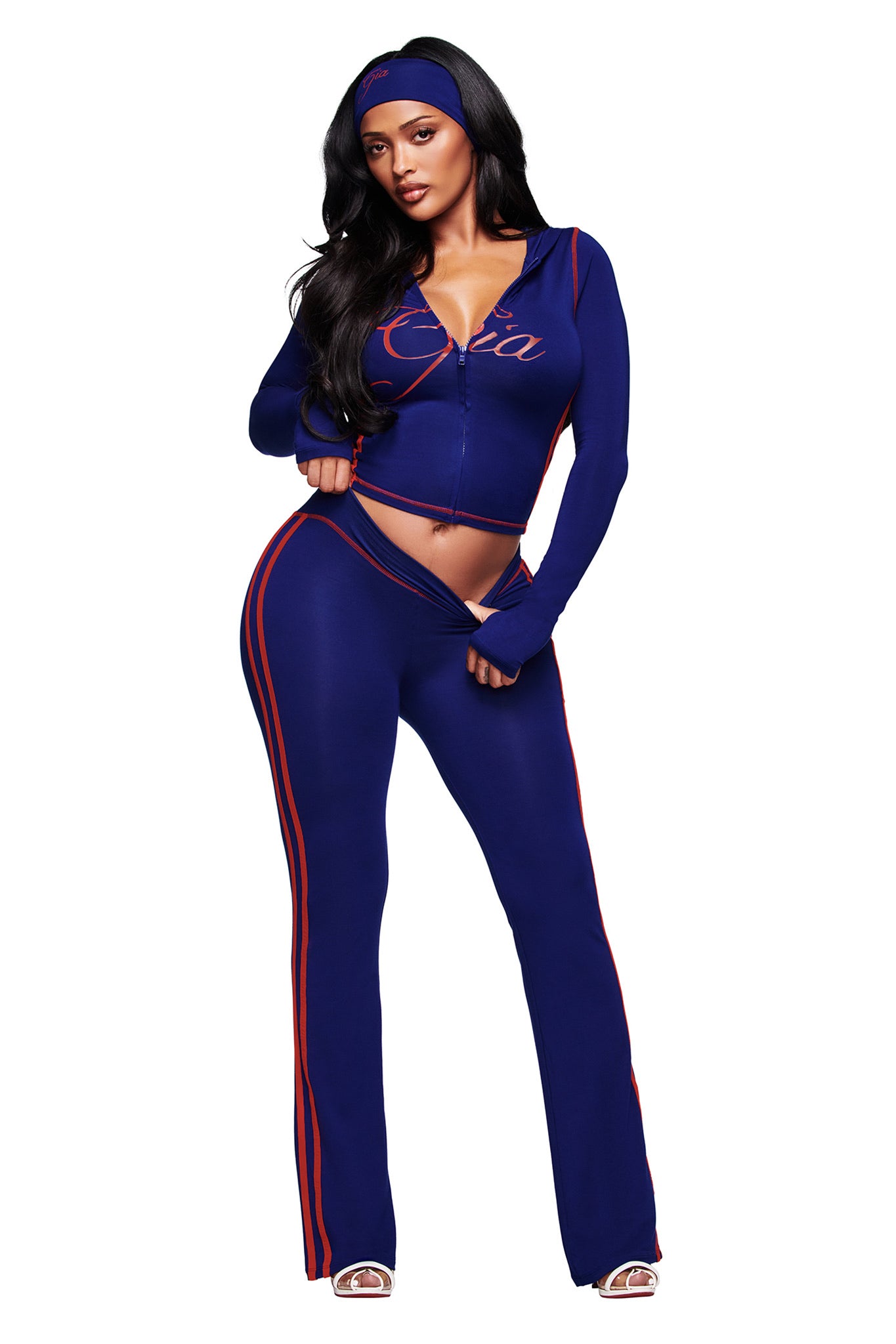 A woman poses confidently in the I.AM.GIA BLARE SPORT HOODIE AND HEADBAND - NAVY, featuring a fitted zip-up jacket, matching pants and headband, with red stripes and buttery-soft fabric against a white background.