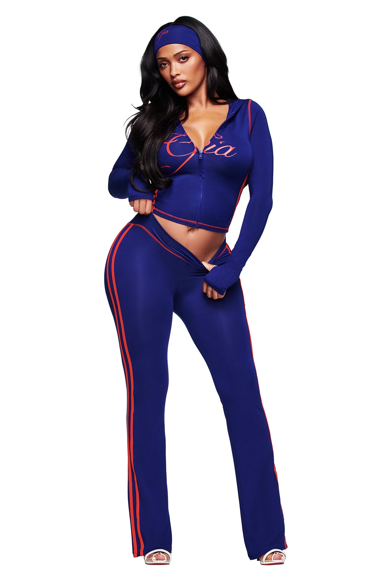 A woman poses in the I.AM.GIA BLARE TWO-PIECE SPORT HOODIE in navy with red stripes and "Coca" on the jacket, styled with a blue headband and open-toe heels against a white background.
