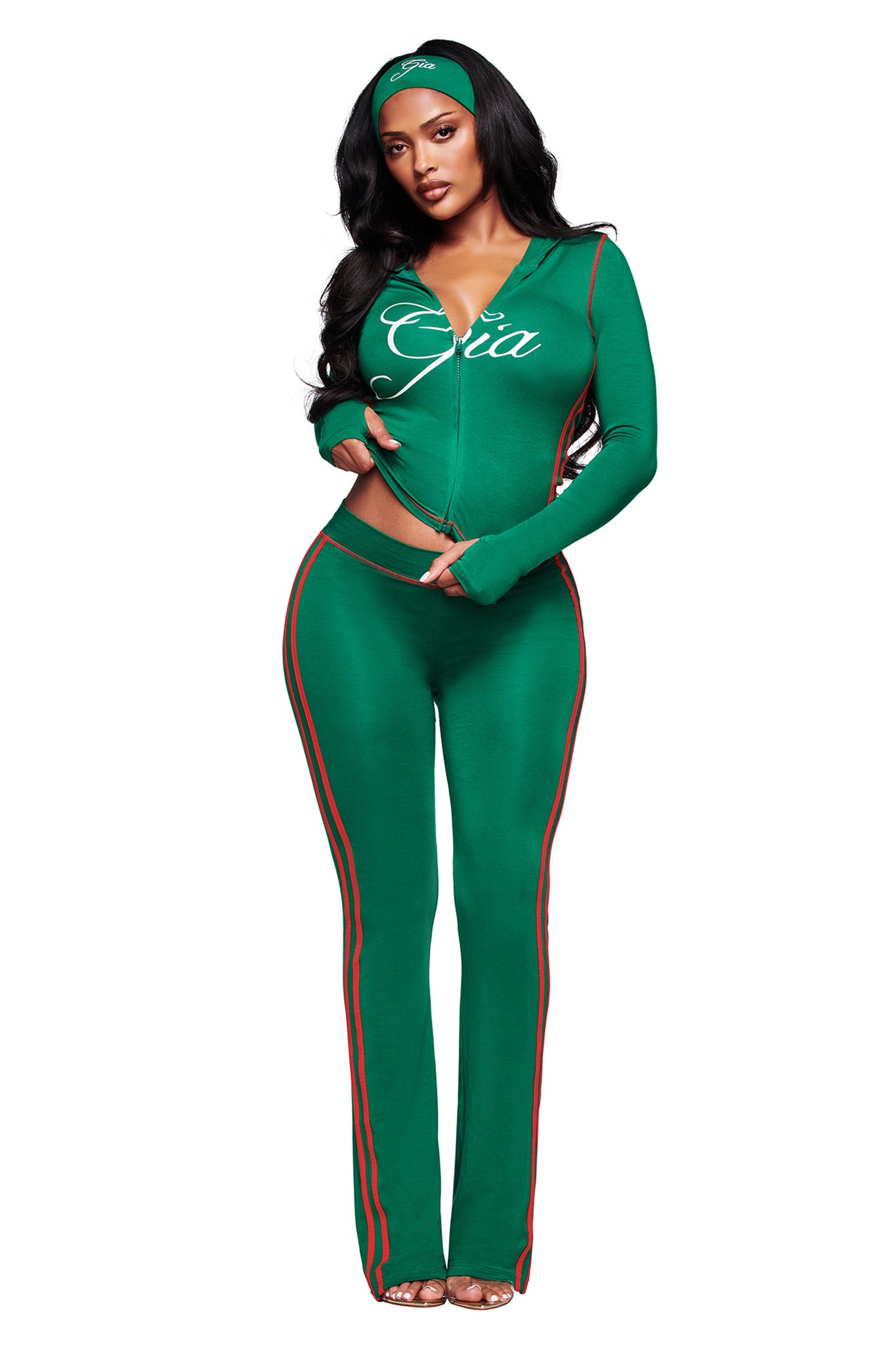 A woman models the I.AM.GIA BLARE TWO-PIECE SPORT HOODIE in green, featuring red stripes and "Gia" on the zip-up top, paired with a matching headband and clear heels, standing confidently against a white background.