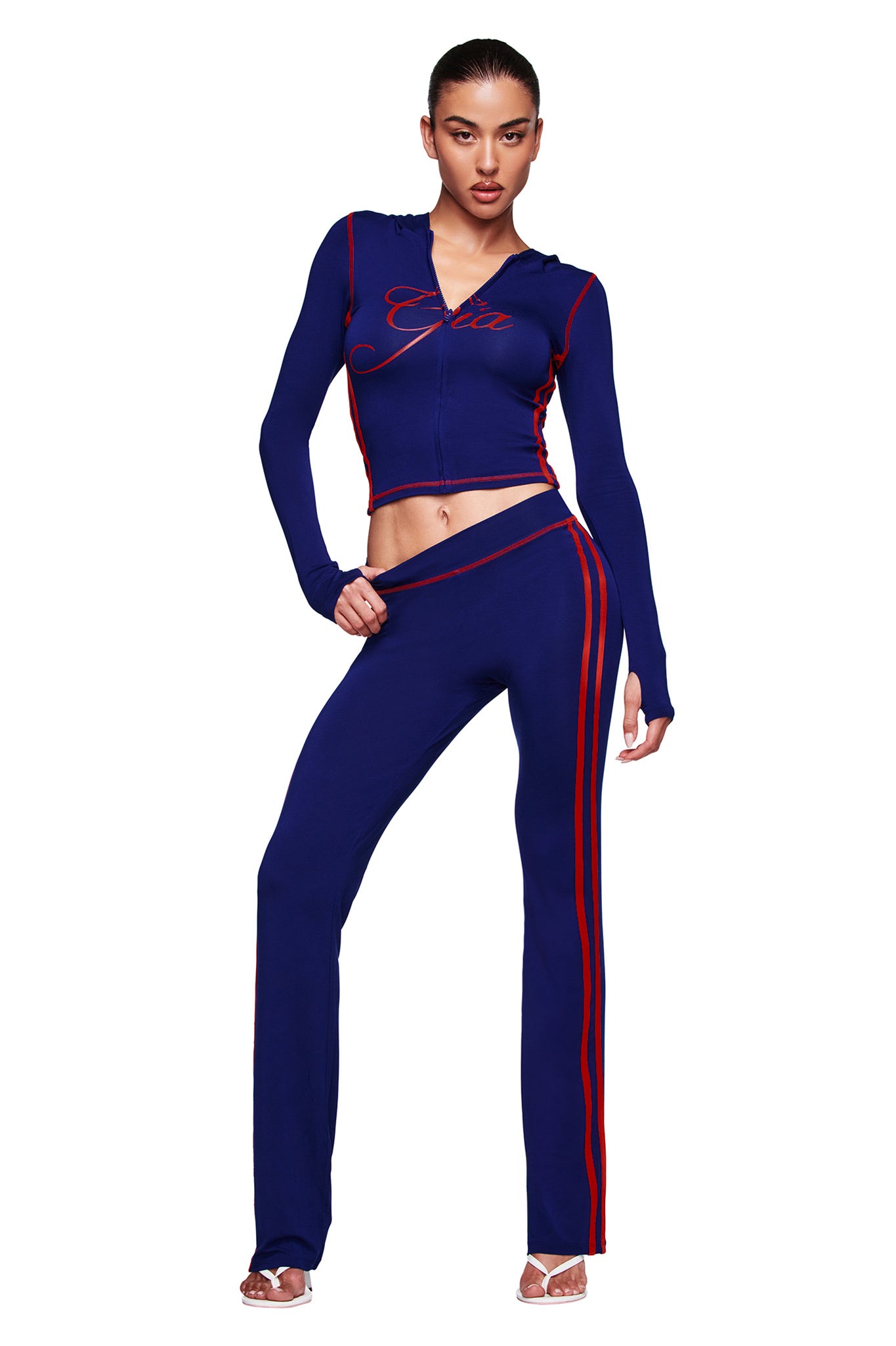 A woman confidently poses in the I.AM.GIA BLARE SPORT HOODIE AND HEADBAND – NAVY, a fitted two-piece tracksuit with red stripes and "Gia" on the zip-up, styled with open-toed heels against a plain white background.