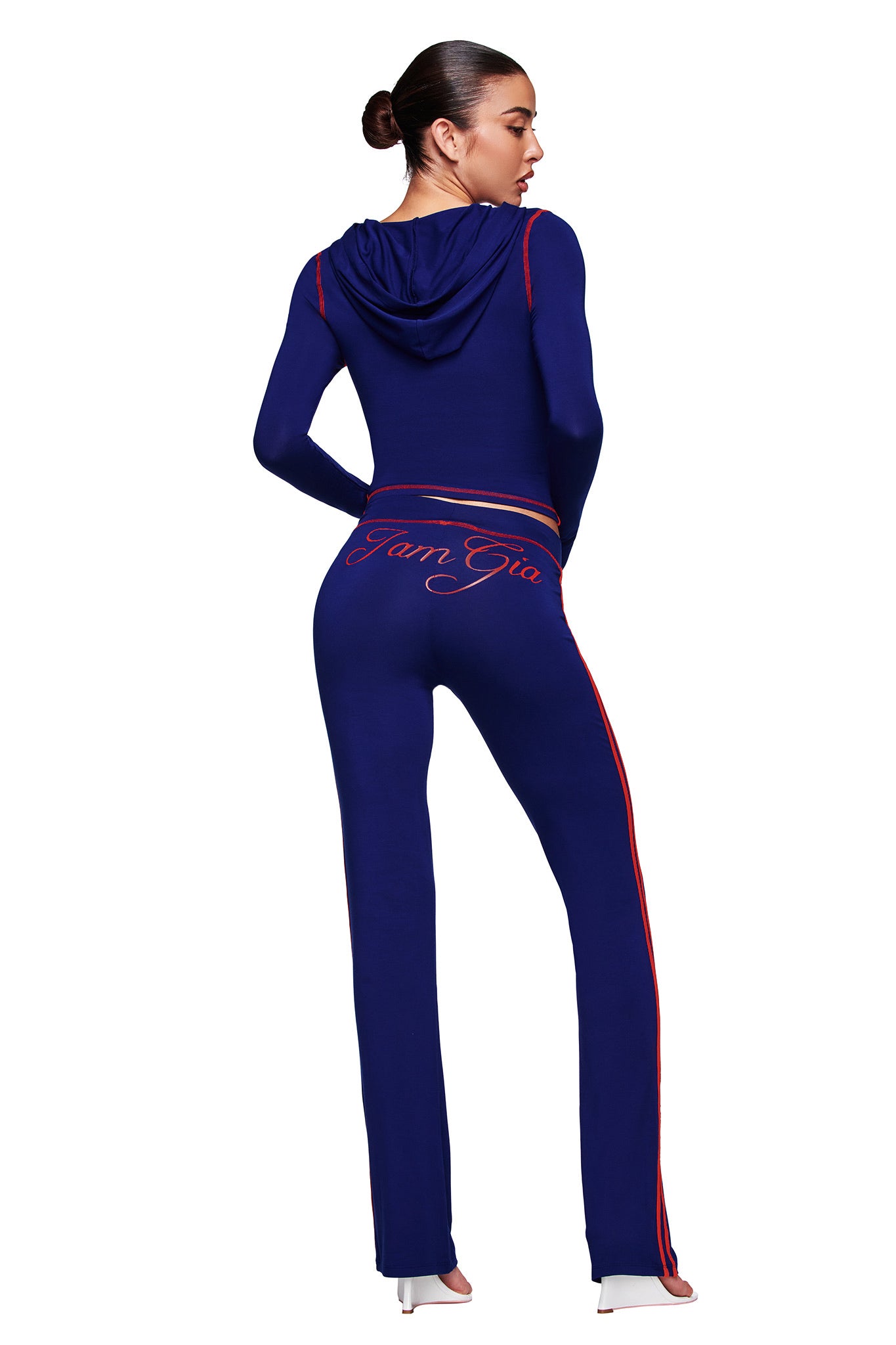 A woman faces away in the I.AM.GIA BLARE Sport Hoodie and Headband set in navy, featuring red trim and a bodycon fit. "I.AM.GIA" script accents her pants, while she styles her hair in a bun and adds white high heels.