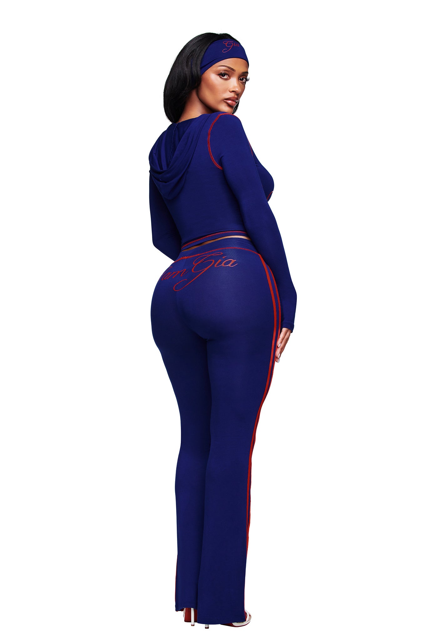 A woman faces away, glancing over her shoulder, wearing the I.AM.GIA BLARE SPORT TRACKPANT in navy with red stripes, a fitted blue long-sleeve top, and a matching headband against a white background.