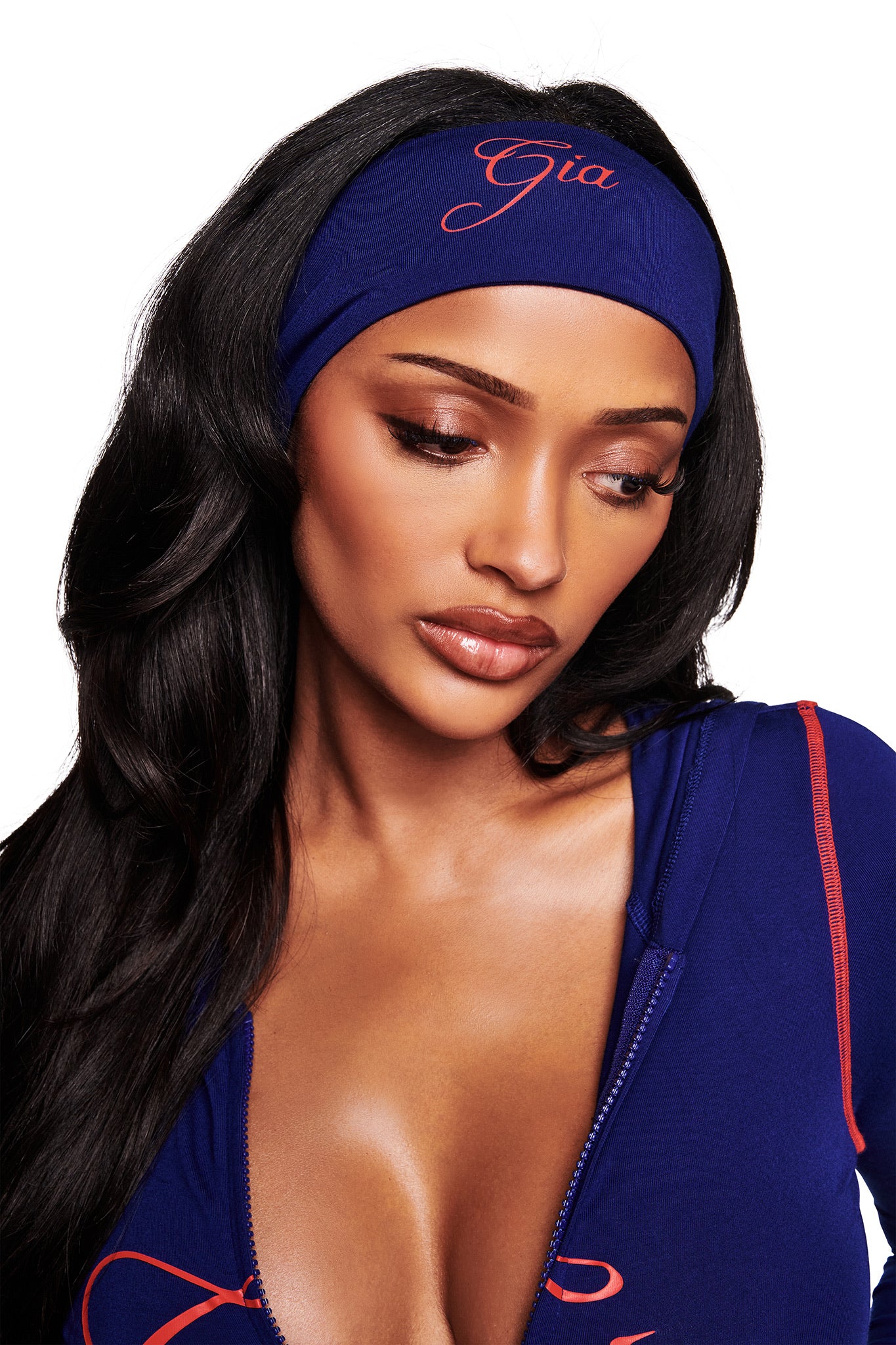 A woman with long dark hair wears the I.AM.GIA BLARE SPORT HEADBAND in navy and a matching blue zipper top with red accents, looking down serenely against a white background.