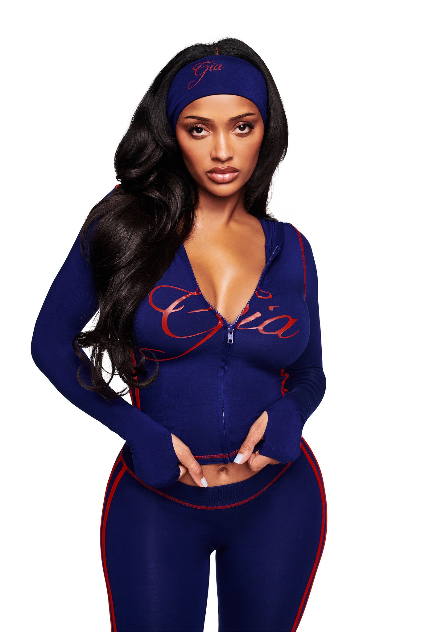 A woman with long dark hair poses against a white background, wearing a navy athletic outfit with red piping and the I.AM.GIA BLARE SPORT HEADBAND - NAVY, as "Gia" appears on both her jacket and sporty headband.