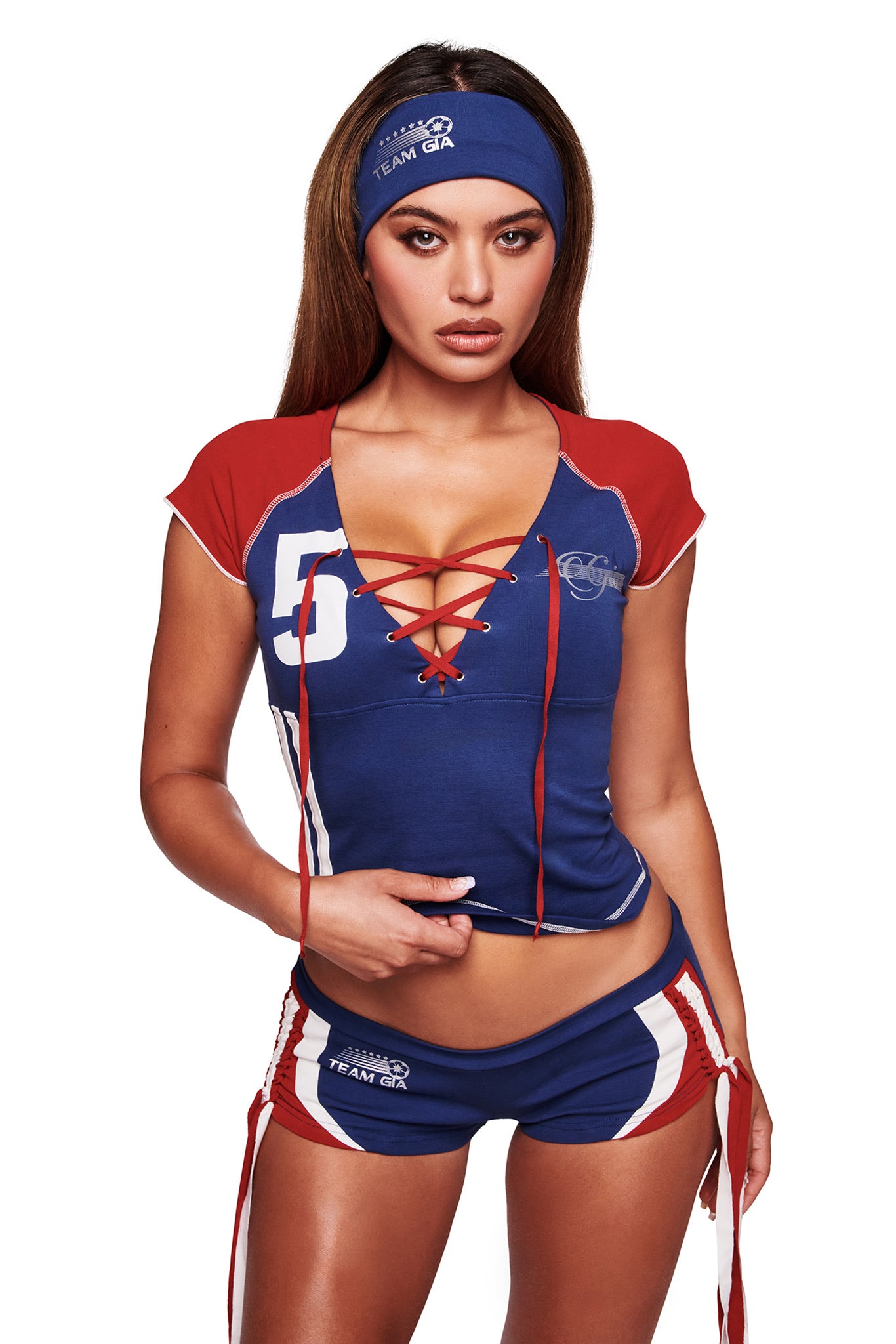 A woman wears the I.AM.GIA NOVIA SPORT HEADBAND in navy with a matching blue athletic set featuring red and white accents, a lace-up front, and the number 5 on the top for a sporty soccer-inspired look against a white background.