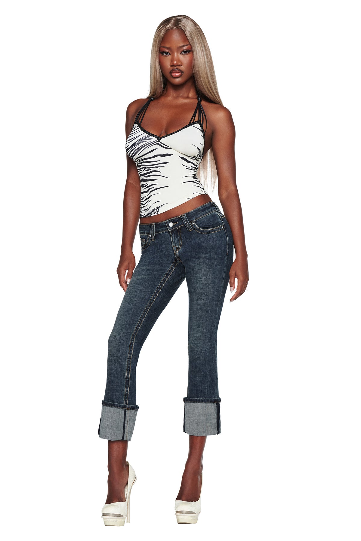 A woman with long blonde hair poses confidently against a white background, wearing a zebra-print tank top, white high heels, and the I.AM.GIA BIANKA JEAN - BLUE: slim straight-leg blue denim jeans rolled up at the cuffs.