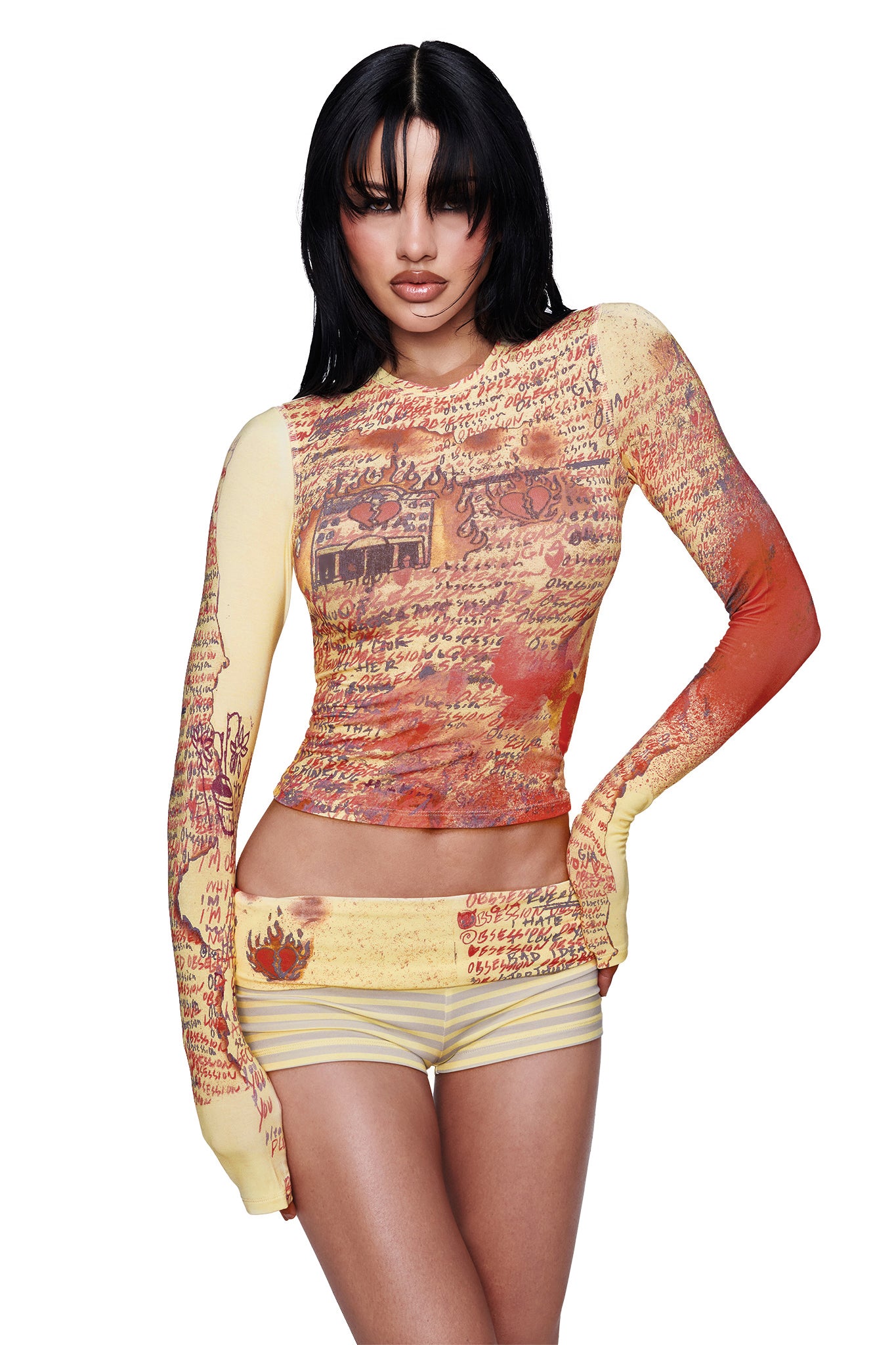 A woman with long black hair and bangs wears the I.AM.GIA T-REX BLARE 2.0 LONG SLEEVE TEE in yellow, a fitted bodycon top featuring vintage-inspired handwritten script and illustration graphics in warm tones, paired with matching shorts, standing against a white background.