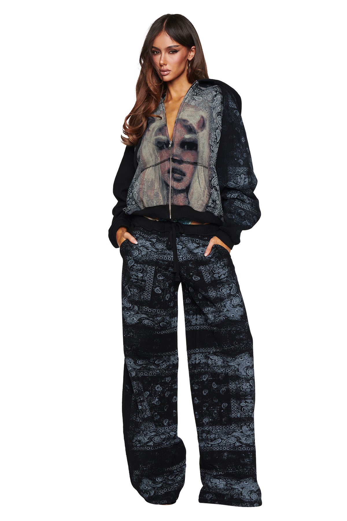 A woman stands with her hands in her pockets against a white background, wearing the I.AM.GIA T-REX ZIP THRU HOODIE in Black Bandana—an oversized street style zip-up hoodie featuring a face graphic—paired with matching black and gray patterned wide-leg pants.