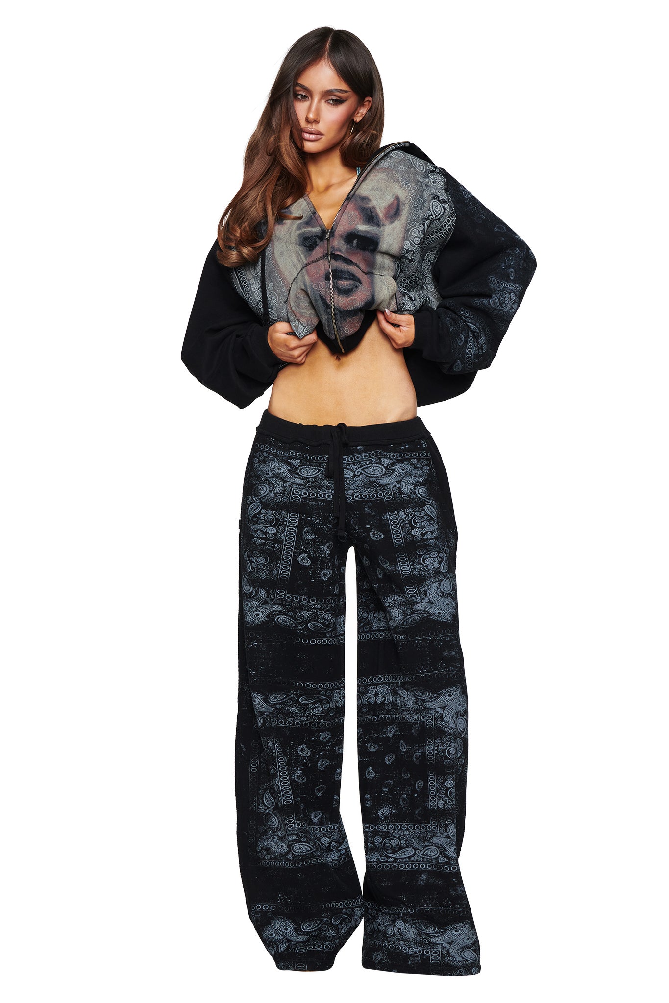 A woman stands against a white background in the I.AM.GIA T-REX TRACKPANT - BLACK BANDANA, pairing the bold black patterned pants with a cropped hoodie that features an oversized face graphic; as she lifts the hoodie to reveal her midriff, she makes a striking contemporary street style statement.