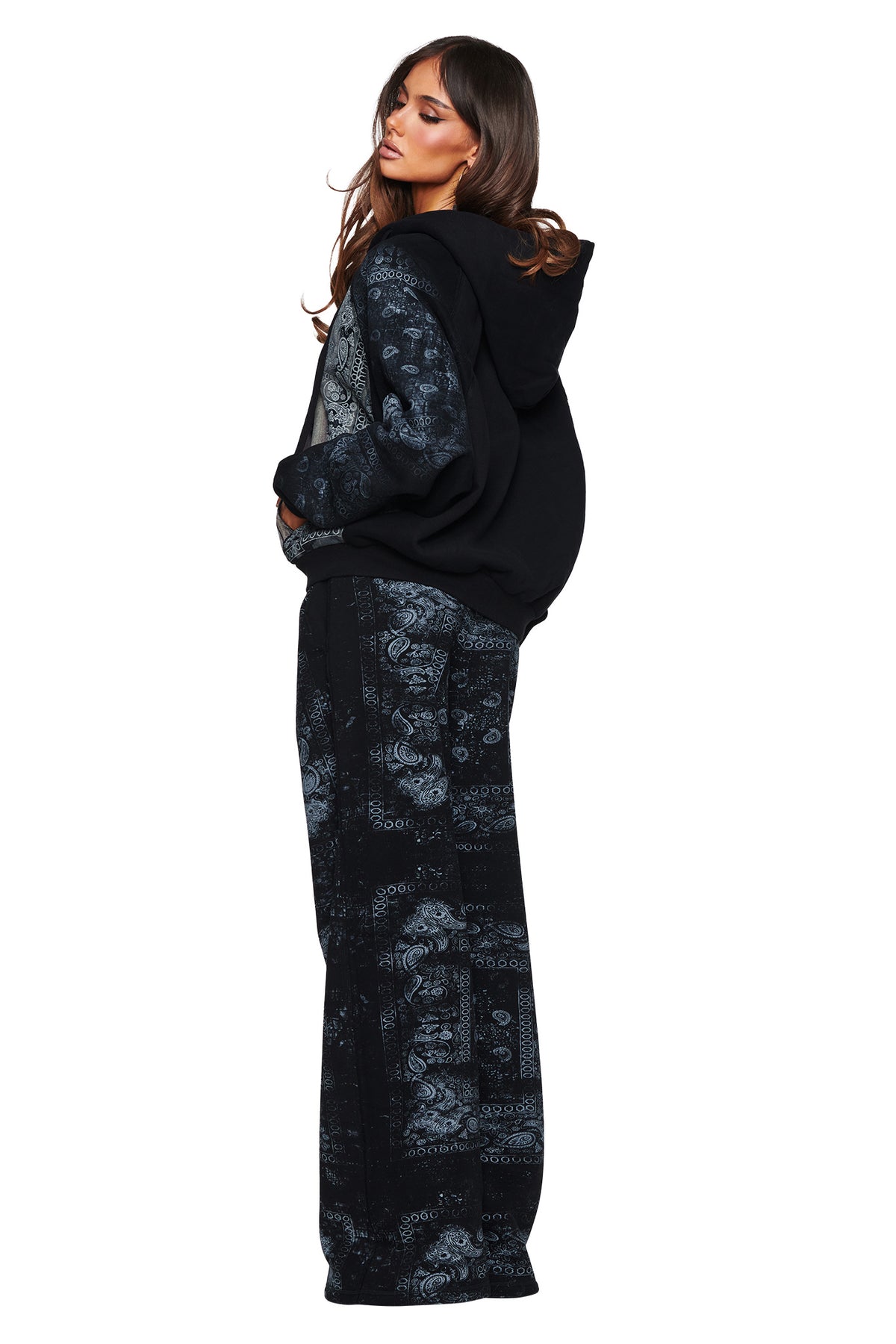A woman stands sideways, wearing the I.AM.GIA T-REX ZIP THRU HOODIE - BLACK BANDANA and matching wide-leg pants, both featuring a gray paisley bandana-style pattern. She looks over her shoulder with long, wavy hair, effortlessly capturing cool street style against a white background.