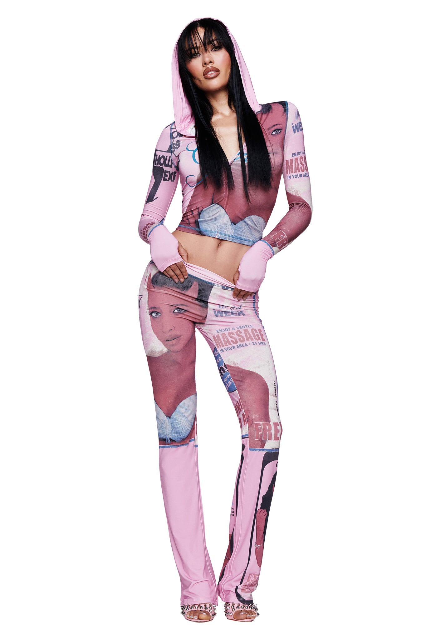 A woman wearing the I.AM.GIA T-REX BLARE HOODIE in pink, featuring magazine-style graphics of a woman's face and upper body, poses with one hand on her hip and the other holding the hood.