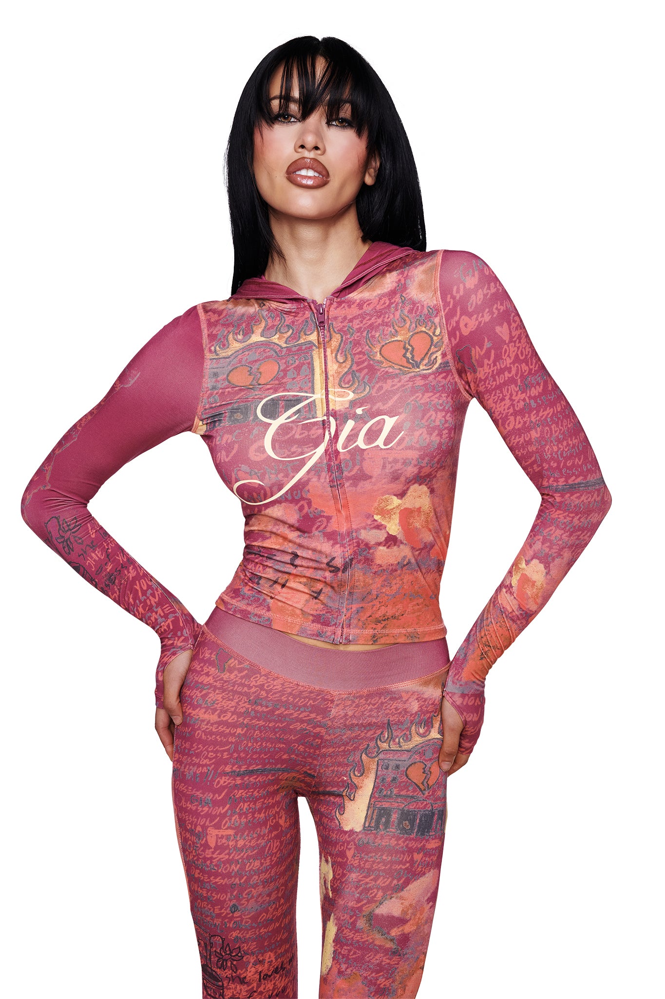A woman with long black hair wears a fitted, colorful patterned I.AM.GIA T-REX BLARE HOODIE in red, featuring "Gia" on the front, as she poses with one hand on her hip against a white background.