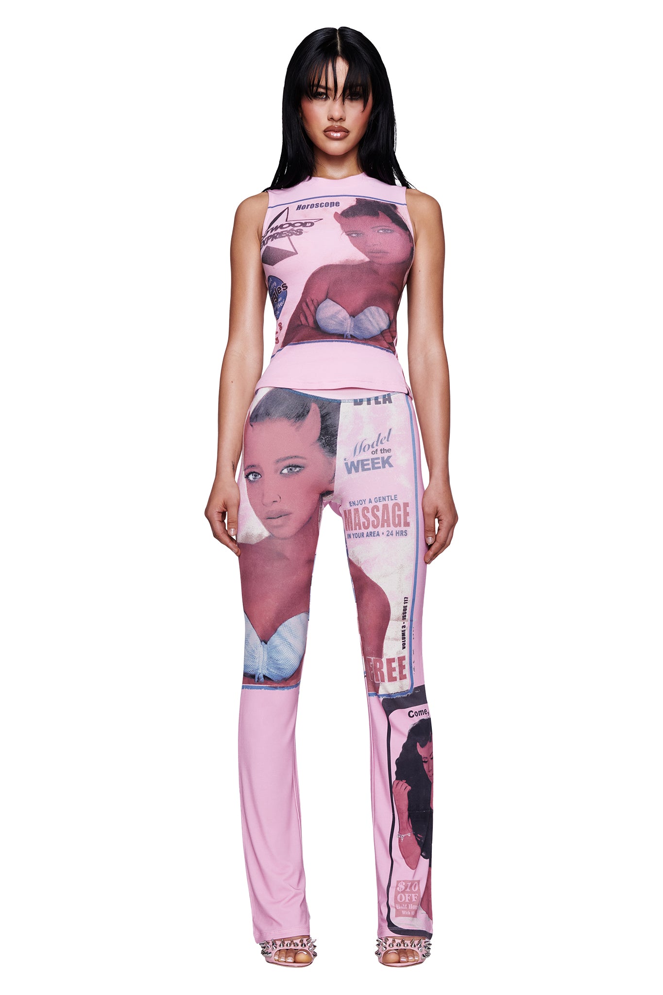 A woman stands facing forward, wearing the I.AM.GIA T-REX BLARE TRACKPANT in pink, featuring an eye-catching T-REX pink print with illustrated female faces and advertising-inspired graphics. The trackpant is fitted with a wide elastic waistband for comfort and style. The background is white.