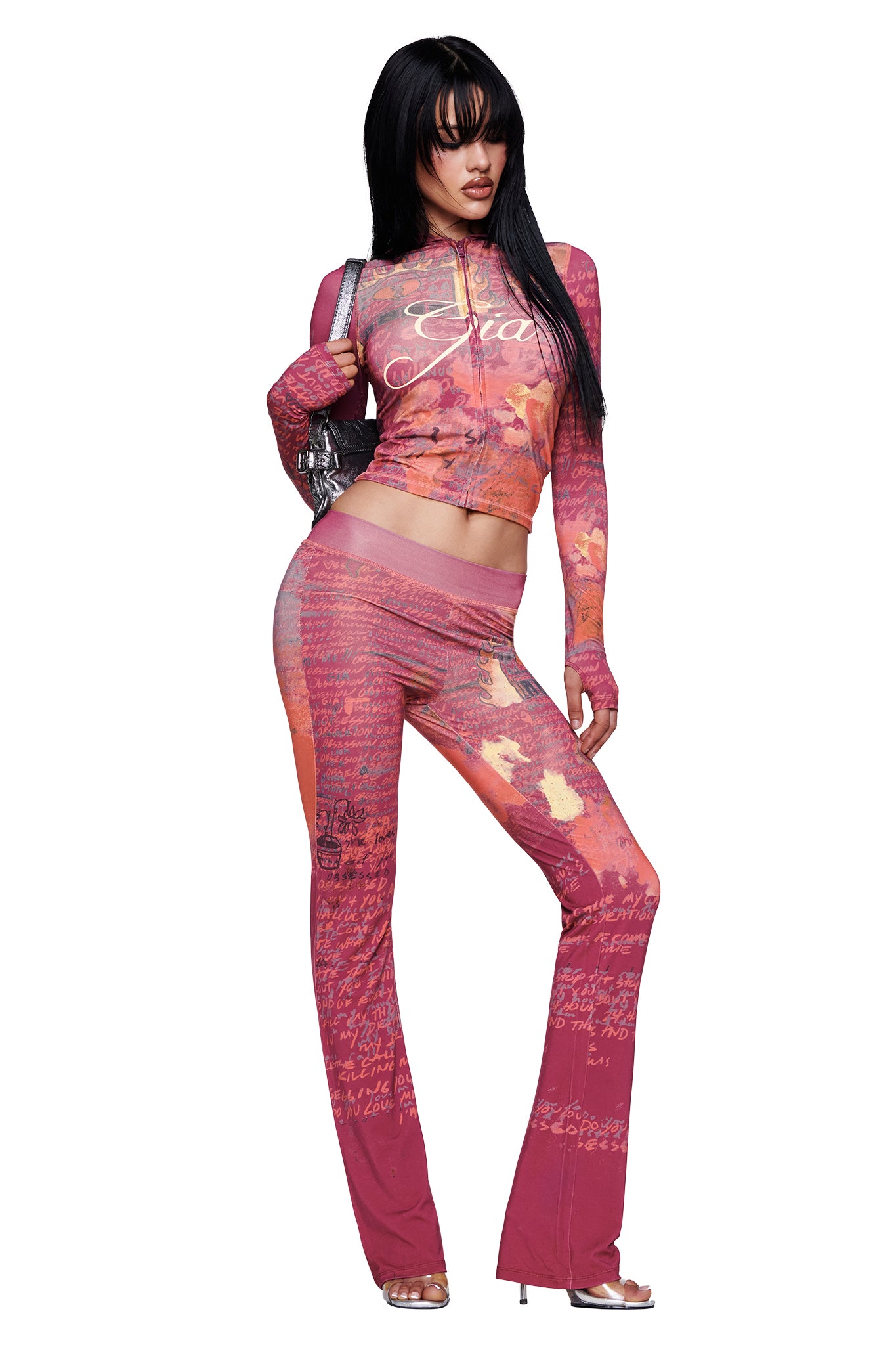 A person poses against a white background wearing the I.AM.GIA T-REX BLARE TRACKPANT in red, styled as part of a matching set with long sleeves. The flared pants feature a wide elastic waistband and are paired with silver heels, a shiny handbag, and long black hair.