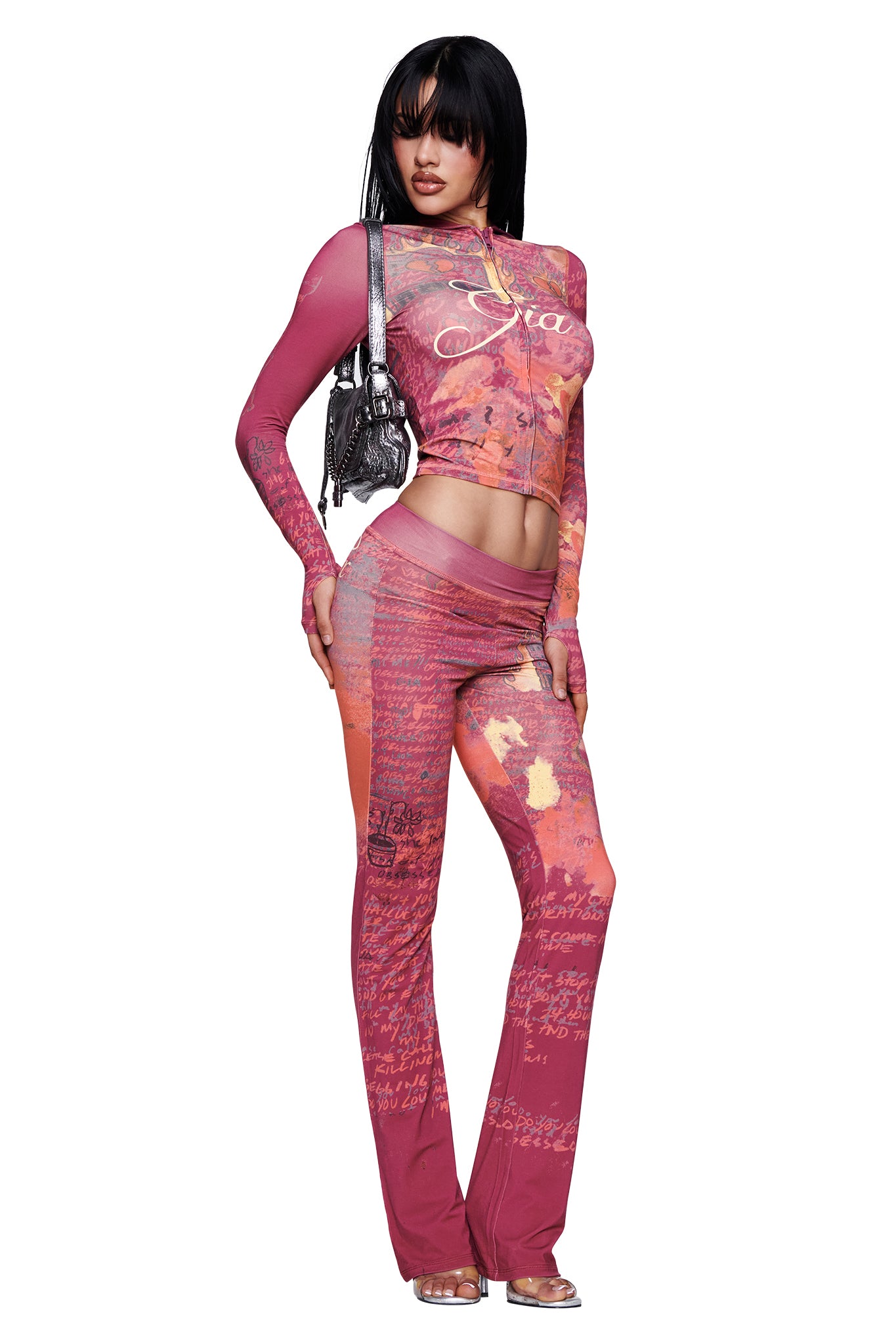 A woman wears the I.AM.GIA T-REX BLARE TRACKPANT in red, styled with a matching fitted long-sleeve crop top featuring T-REX red print and bold paint-effect patterns. The set includes a wide elastic waistband for comfort. She accessorizes with a small silver purse and clear high-heeled sandals.