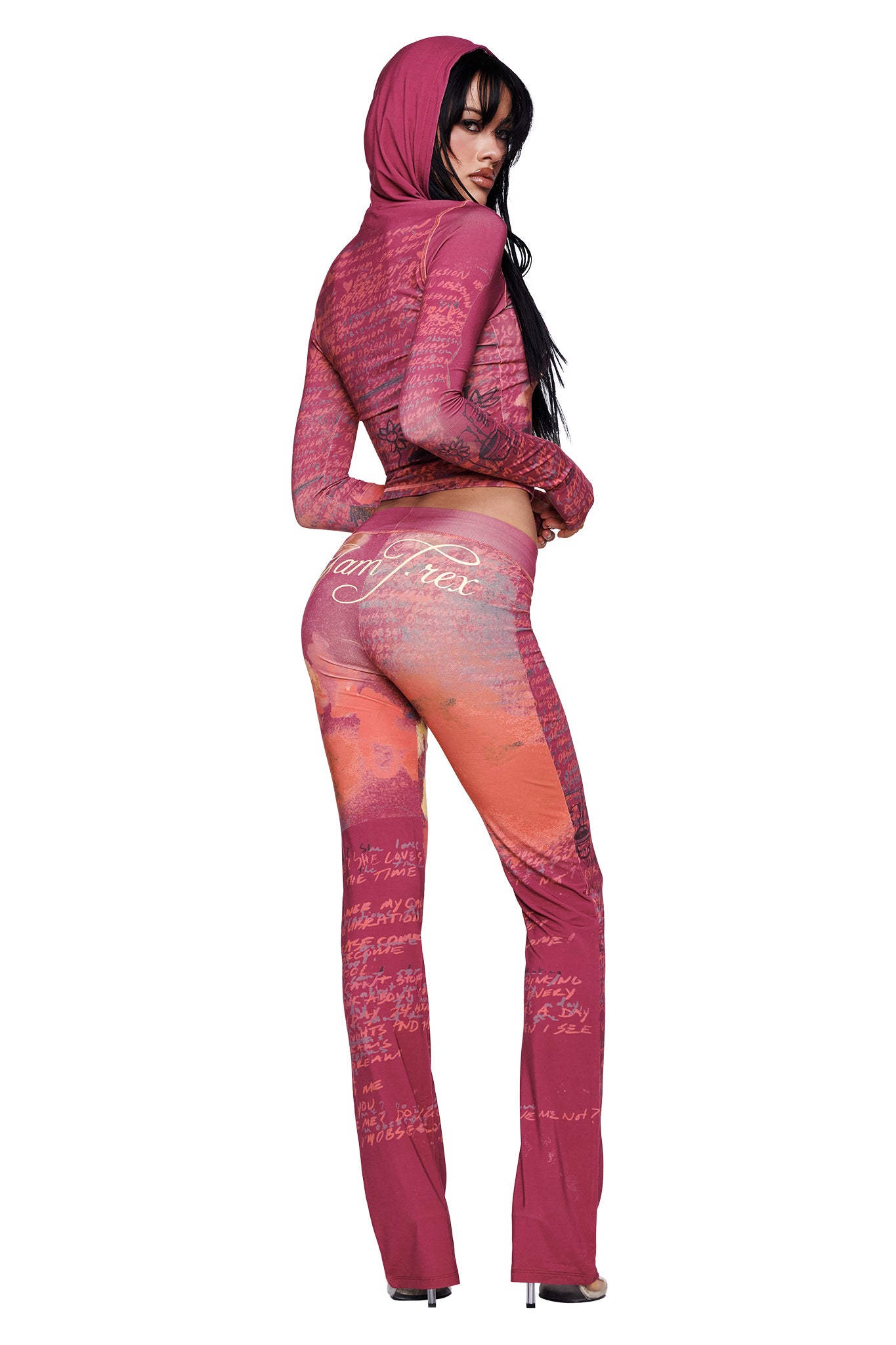 A woman stands in high heels, wearing the I.AM.GIA T-REX BLARE HOODIE in red—a fitted, hooded outfit with script writing and patterns—posing with her back slightly turned and looking over her shoulder.