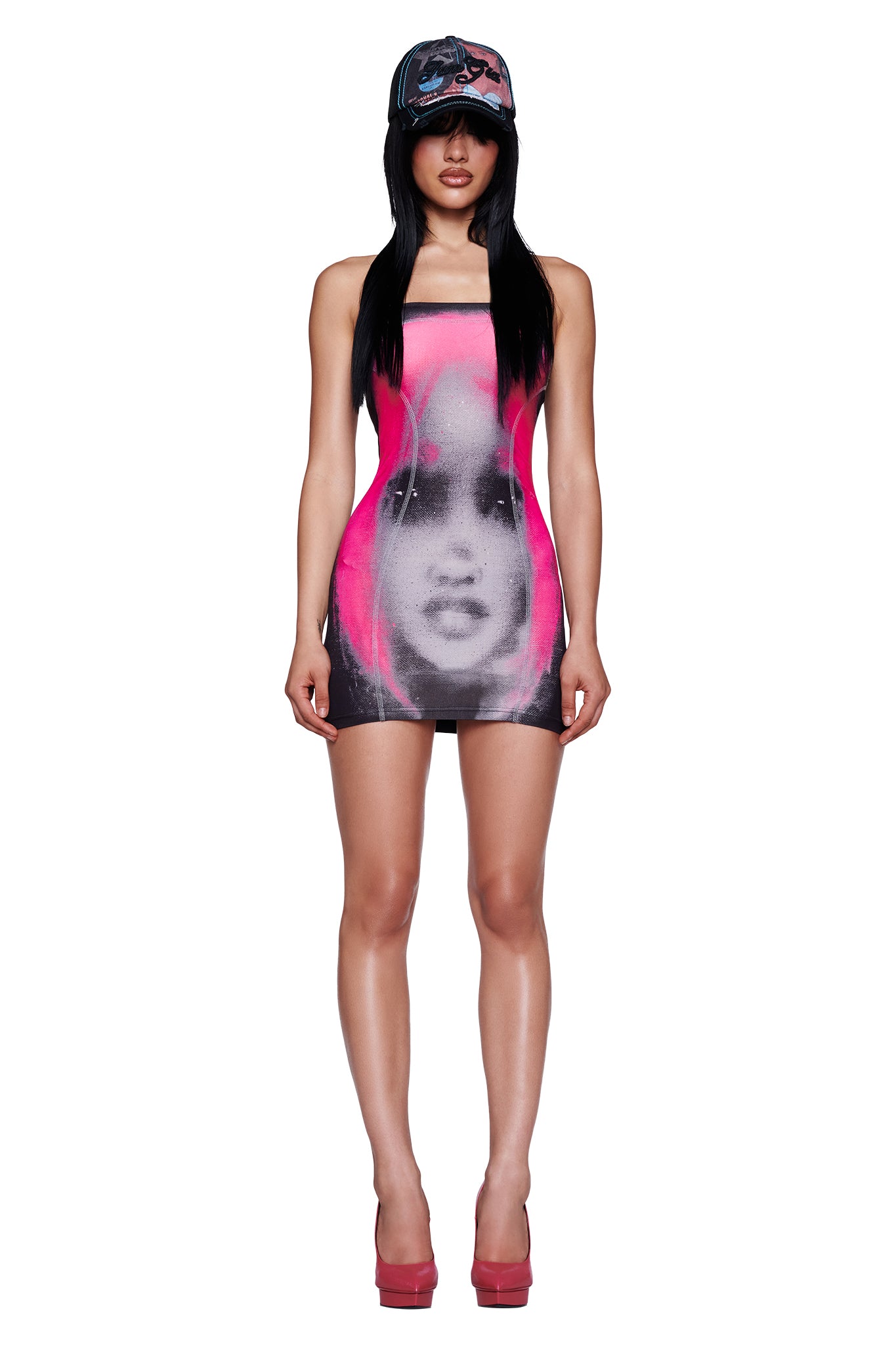 A woman stands in the I.AM.GIA T-REX BLARE BOOB TUBE MINI DRESS in black, featuring a striking pink and black artistic face print, styled with high heels and a baseball cap against a white background.